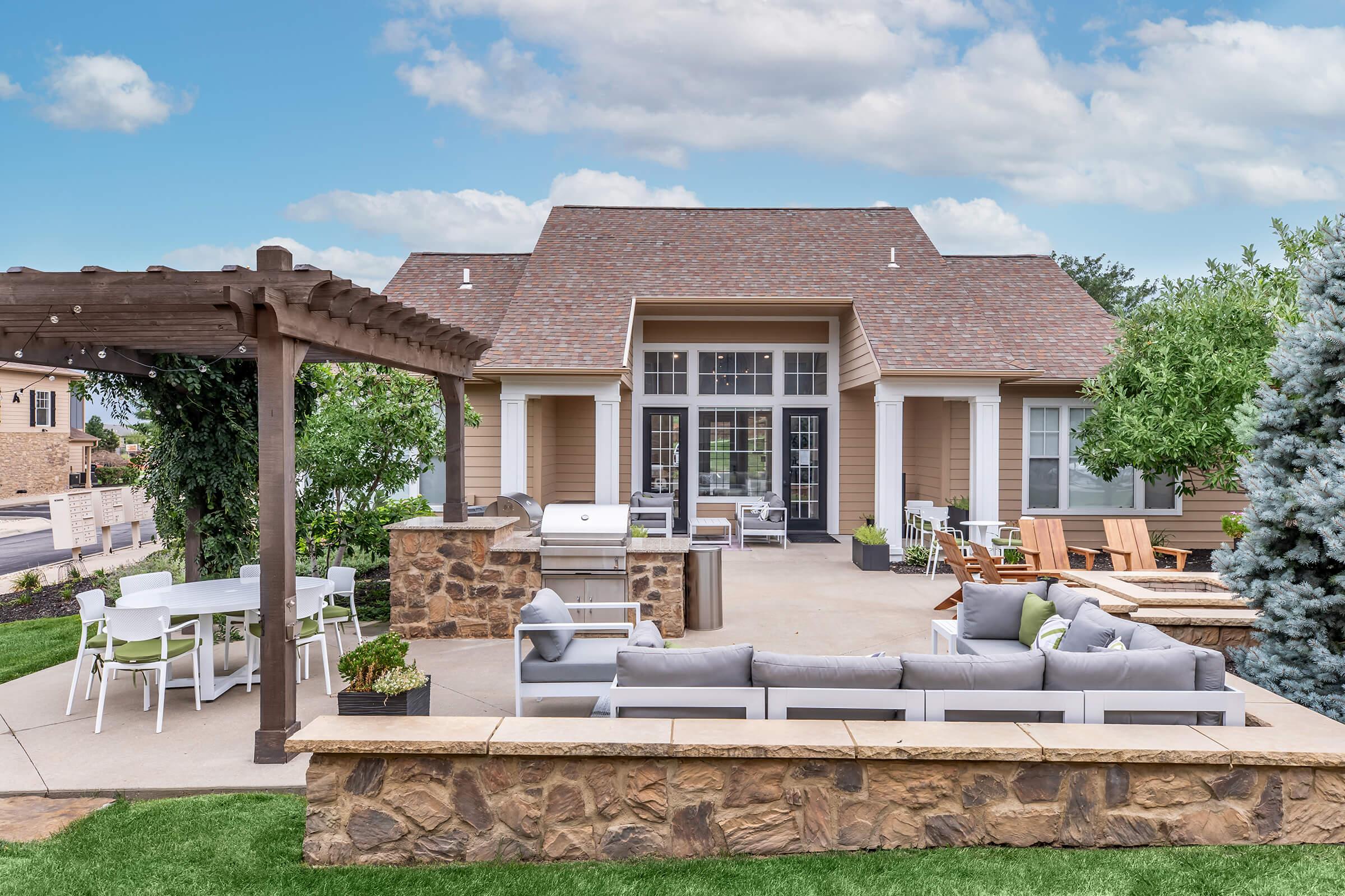 A modern outdoor patio area featuring a large stone wall, comfortable seating with cushions, a dining table, and a grill. The house in the background has a gabled roof, large windows, and a light color scheme, surrounded by trees and a blue sky.