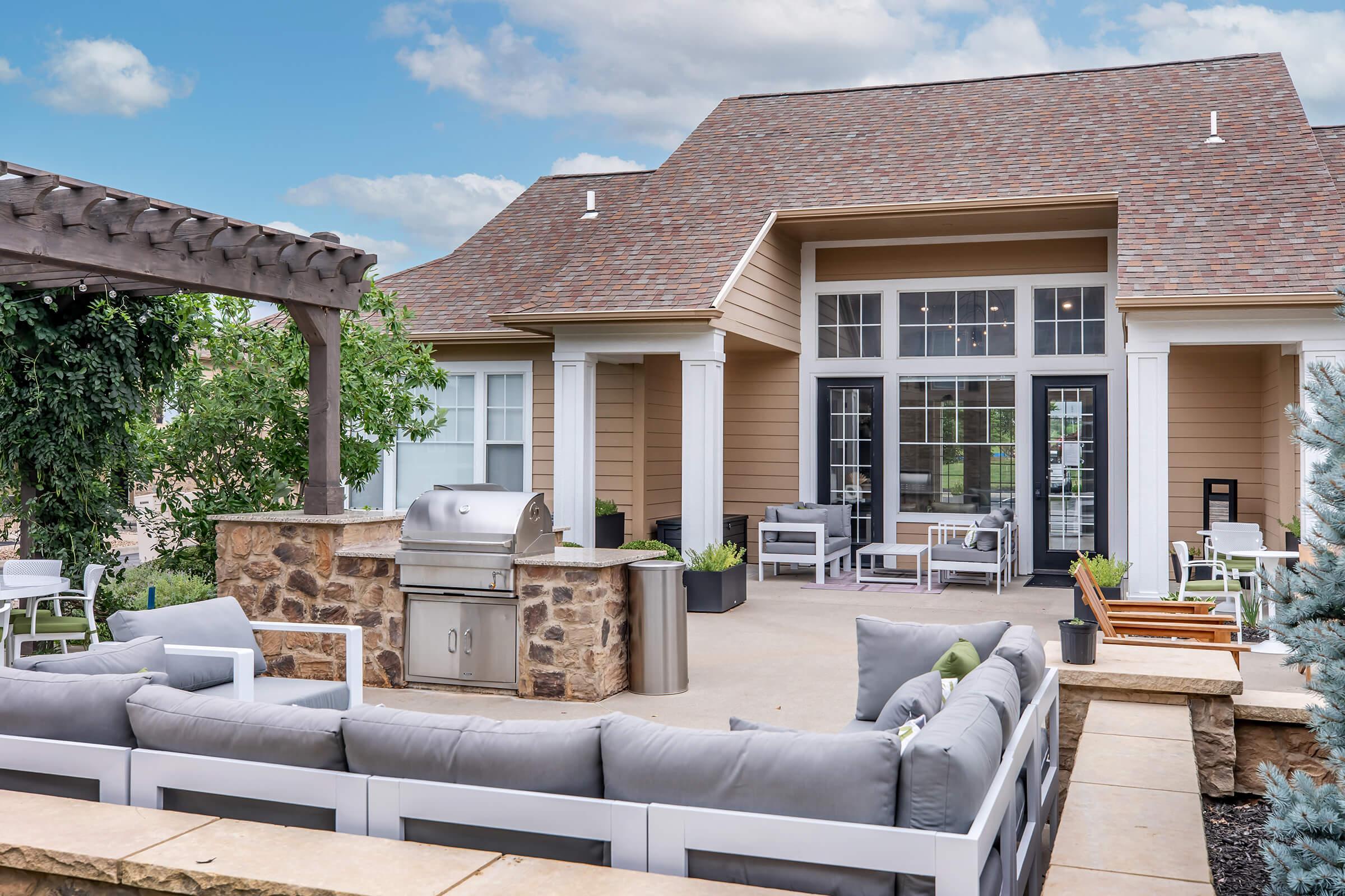 Outdoor patio area featuring a modern grill, stone countertop, and comfortable seating with gray cushions. The setting includes a nearby pergola and greenery, with a backdrop of a stylish house with large windows and a brown shingled roof against a partly cloudy sky.