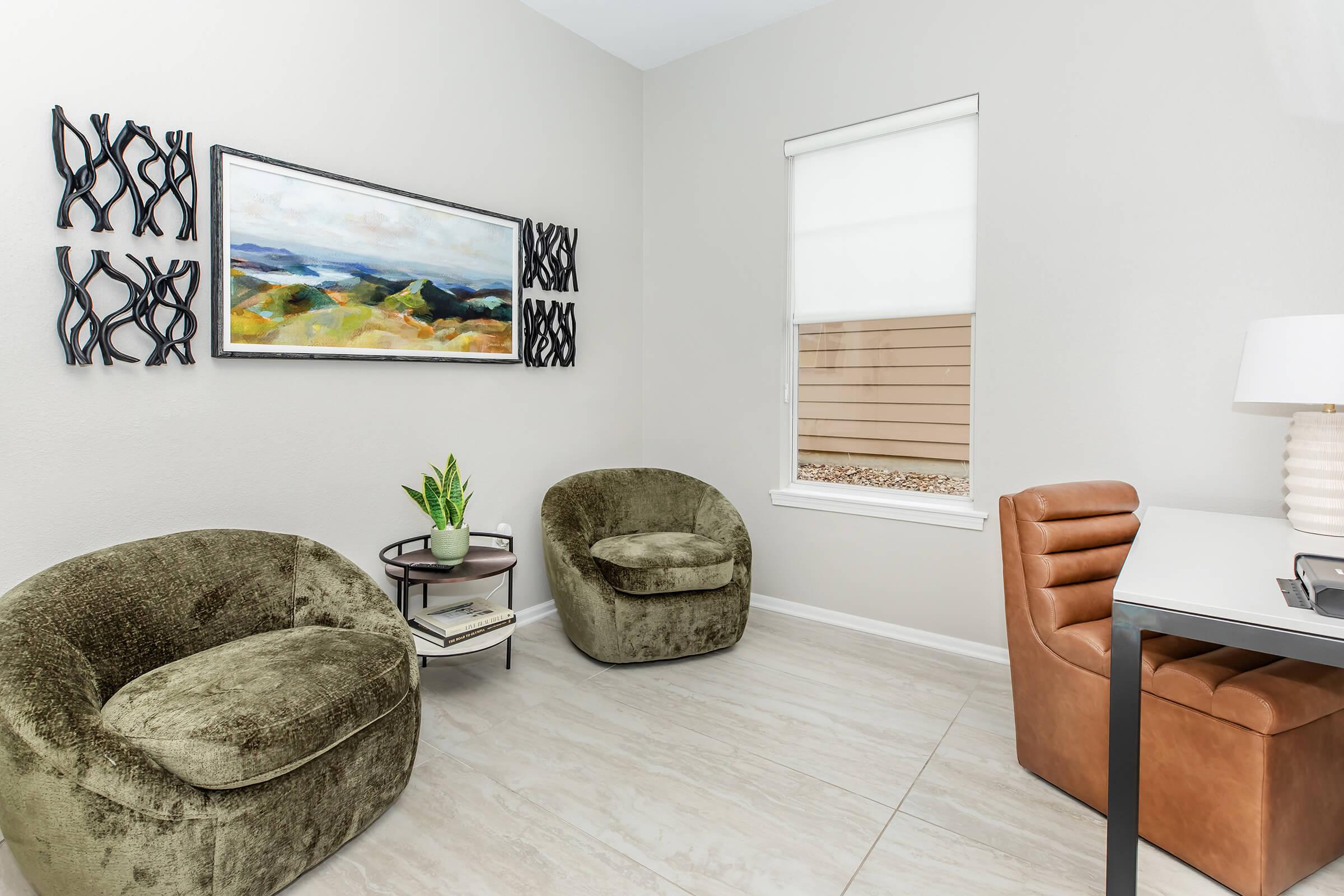 A cozy office corner featuring two green plush chairs, a small round table with a plant, and a stylish desk with a lamp. Above the chairs hangs a colorful abstract landscape painting, and a window with a white blind shows a view of wooden siding outside. The floor has light-colored tiles.