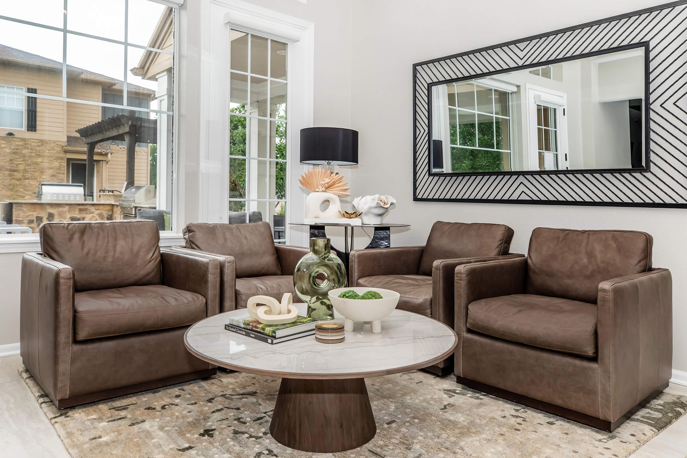 A modern living room featuring two brown leather armchairs, a round marble-topped coffee table, and a decorative centerpiece. A large mirror with a geometric design hangs on the wall, reflecting natural light from the windows. The background includes greenery outside and decorative items on the table.