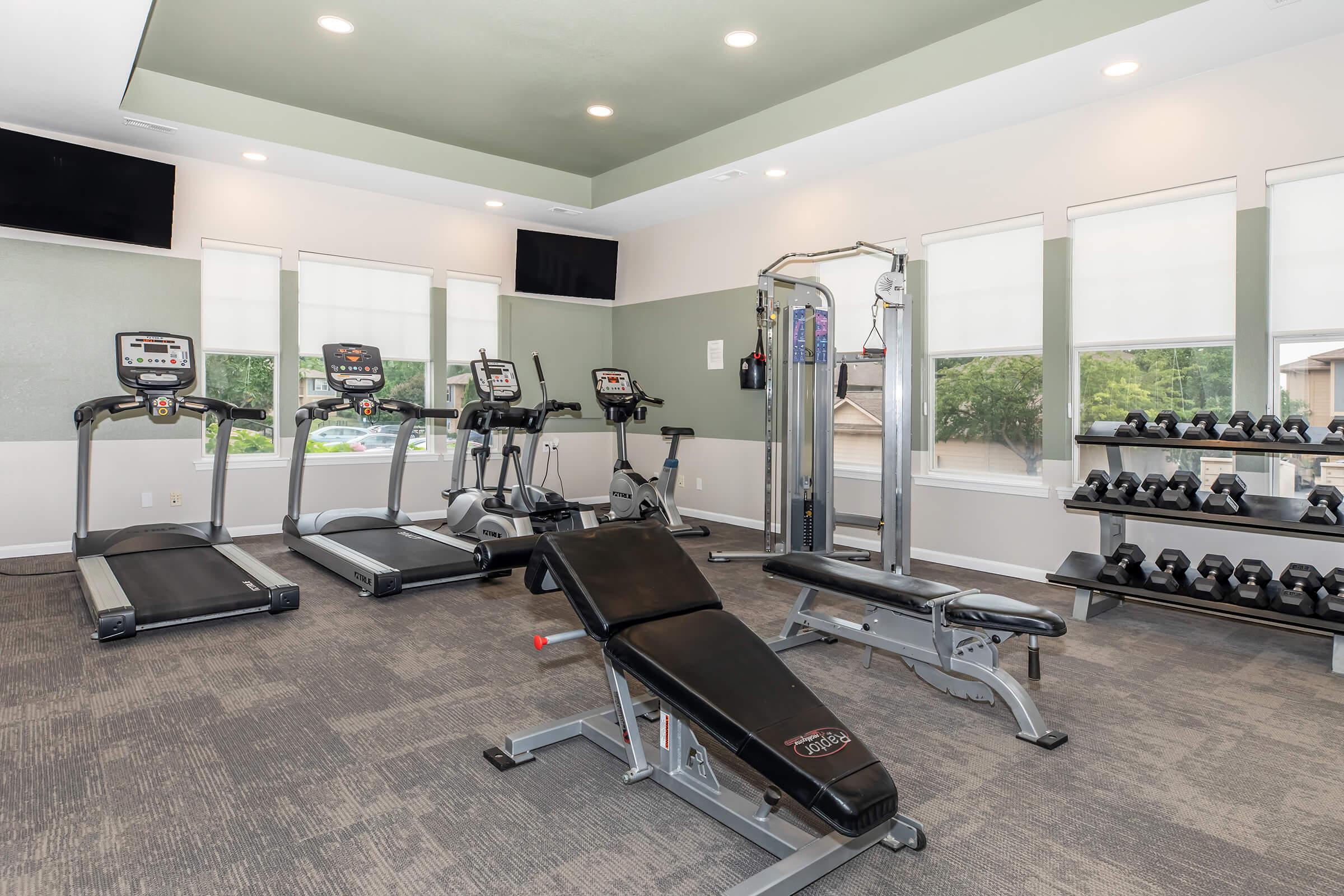A modern fitness room featuring several workout machines, including treadmills and an elliptical. There is a multi-use weight machine, a flat bench, and a set of dumbbells on a rack. Large windows allow natural light to fill the space, creating a bright and inviting atmosphere for exercise.