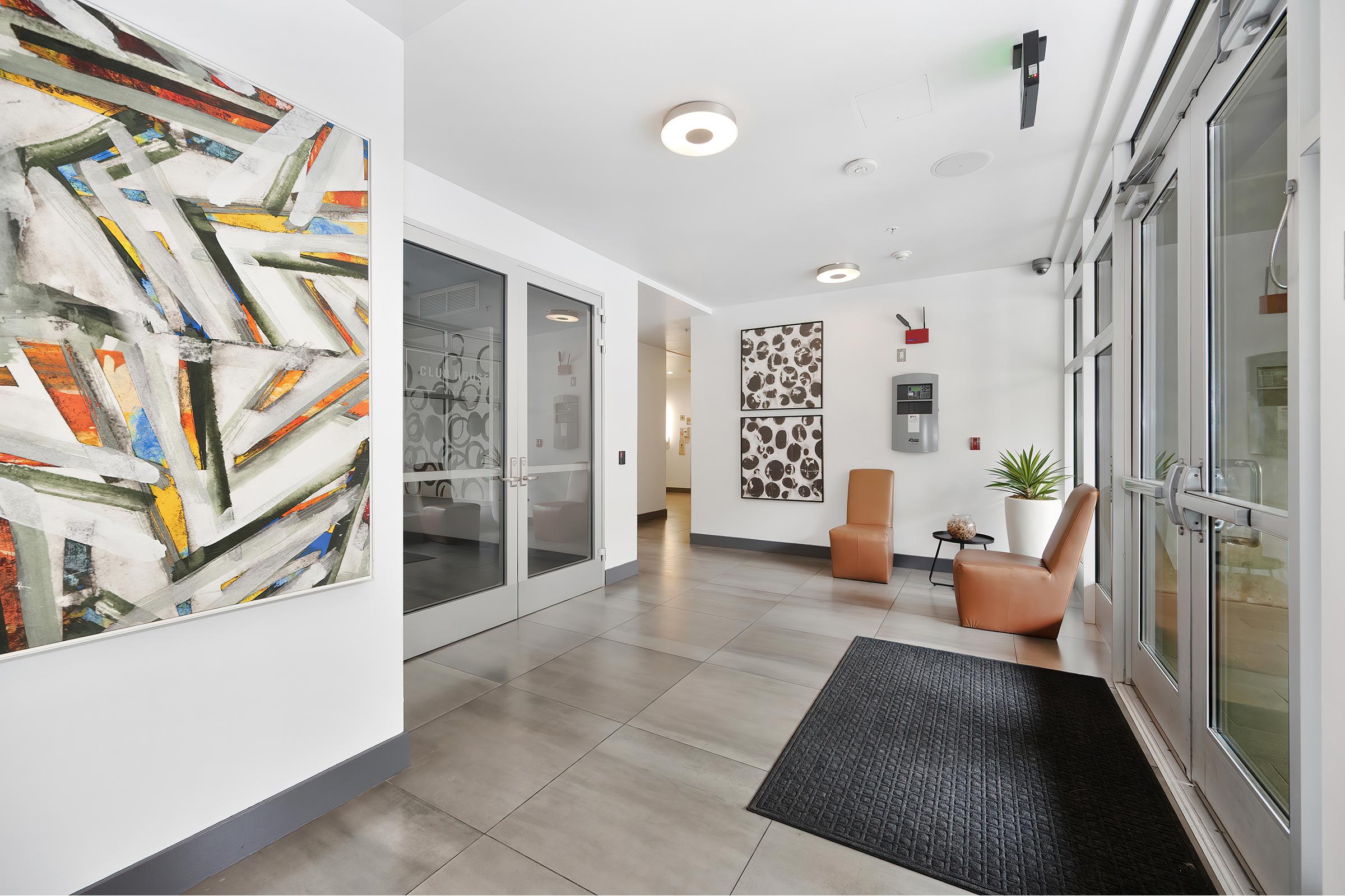 A modern entrance hallway featuring large glass doors, two stylish orange chairs, and a textured gray floor. The walls are adorned with abstract artwork, including colorful geometric designs and patterned pieces. A small plant adds a touch of greenery to the space.