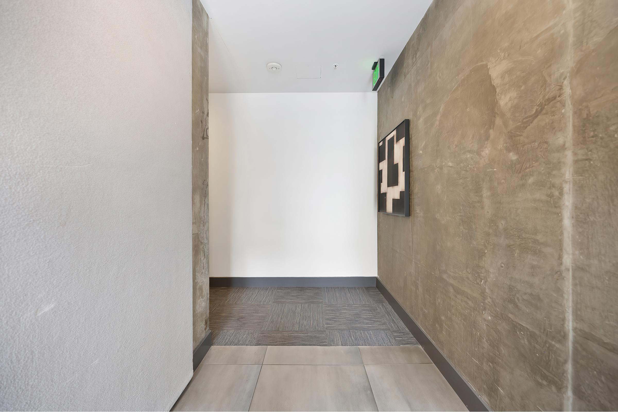 A well-lit hallway with a modern design, featuring smooth concrete walls and a tiled floor. At the end of the hallway, there is a white wall with a framed artwork displayed. The space appears minimalist and inviting, with natural light enhancing the clean aesthetics.