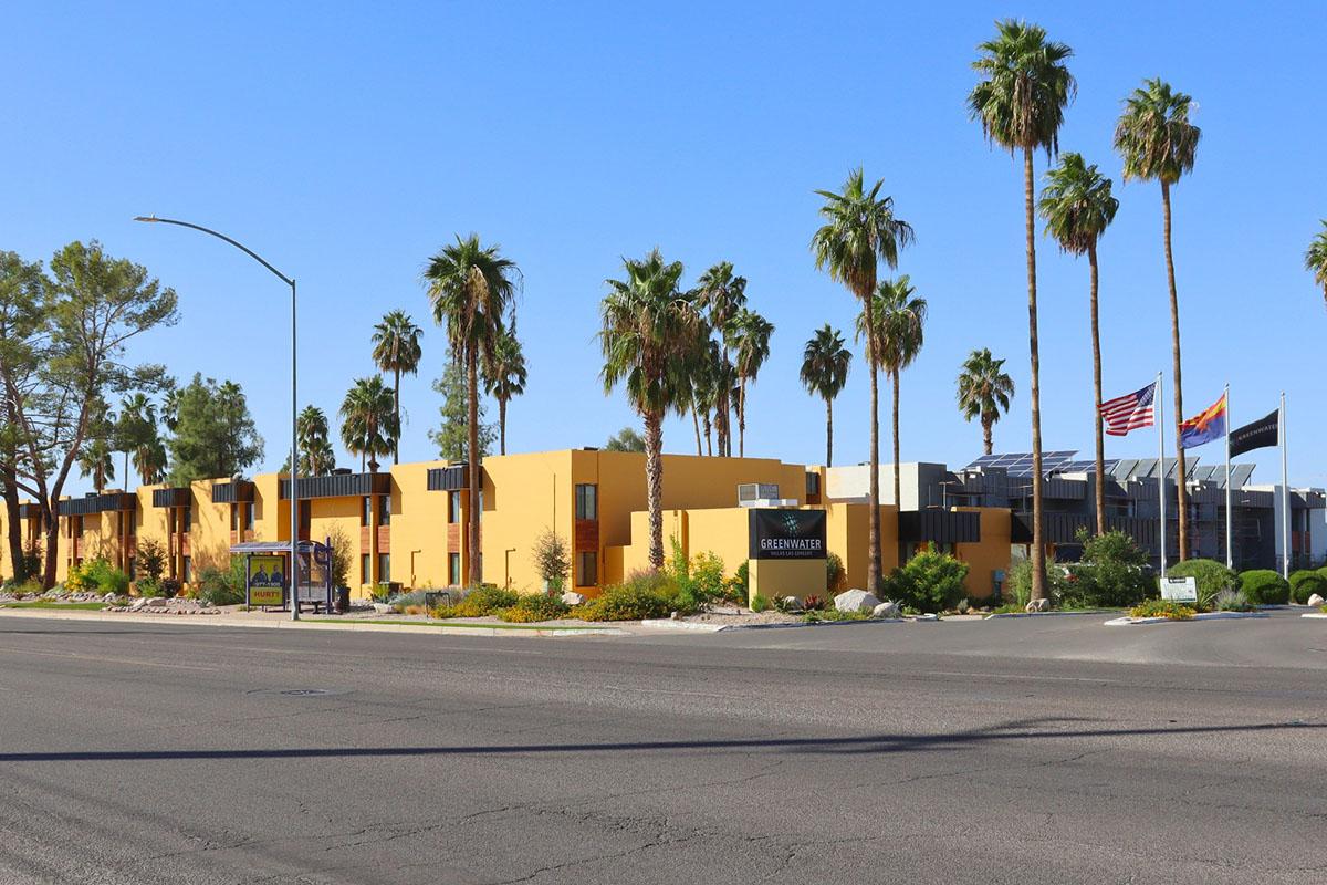 A yellow building with multiple windows is lined with tall palm trees. In the foreground, a street runs alongside the property, with a sign indicating the building name. There are flags of the United States and Arizona visible near the entrance, set against a clear blue sky.