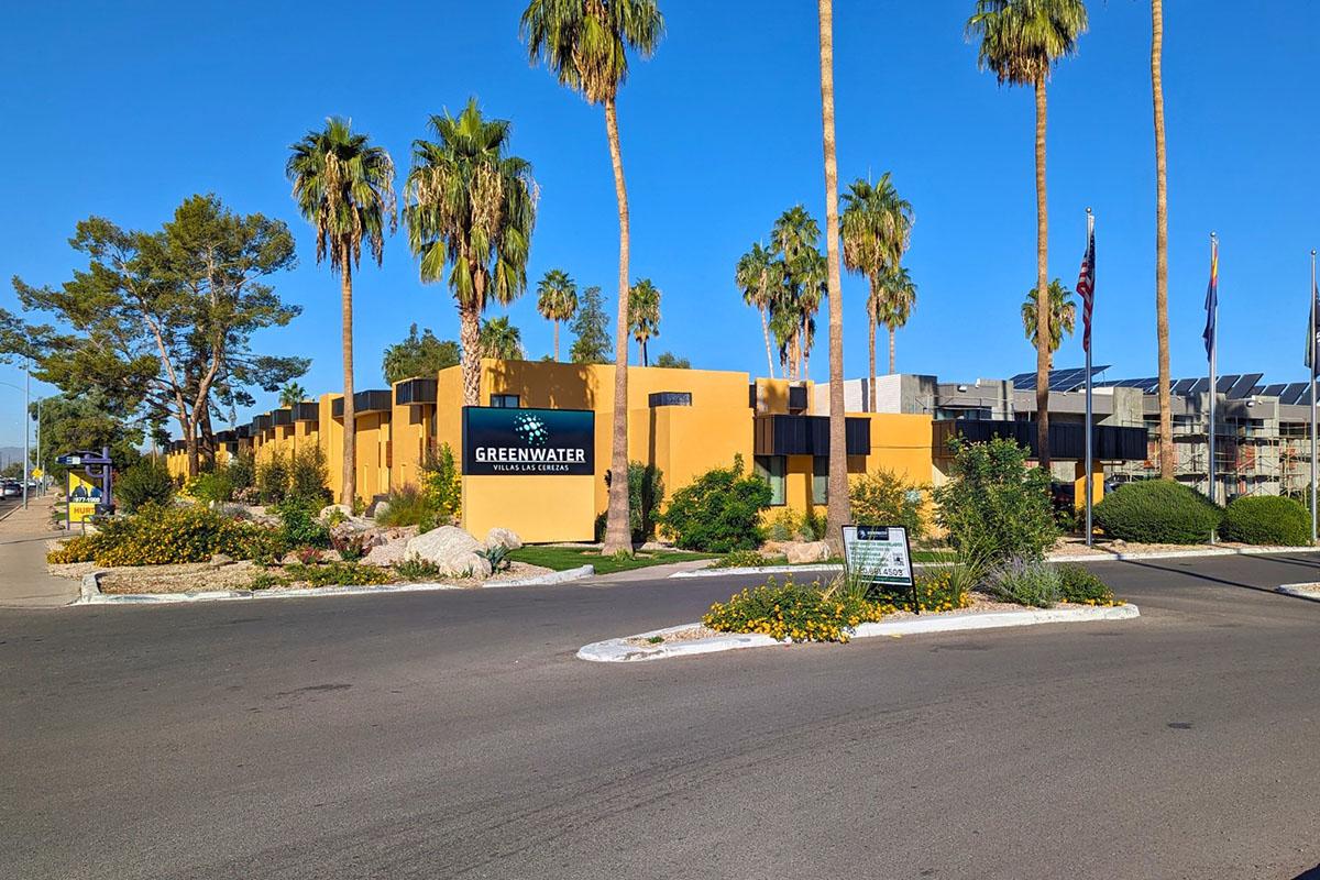 A brightly colored building with yellow walls, surrounded by palm trees and well-maintained landscaping. The sign reads "Greenwater" at the entrance, and there are flags in the background. The scene is set on a clear day under a blue sky.