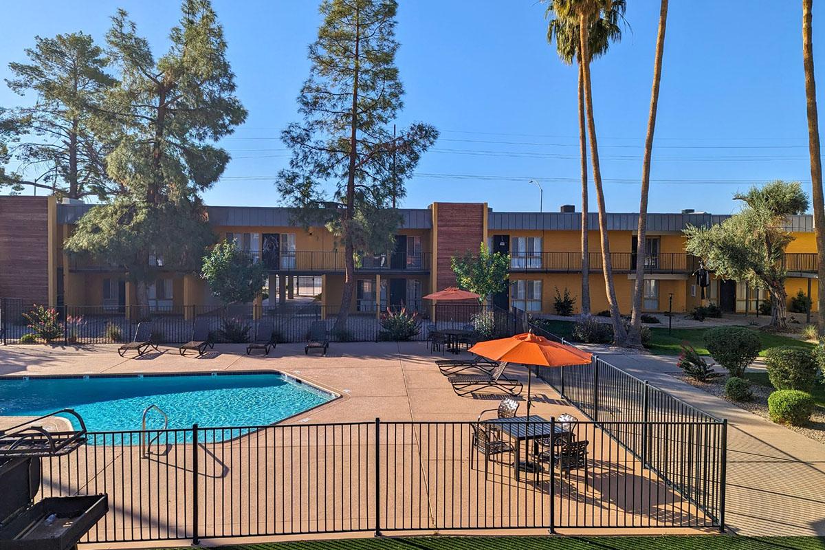 A sunny view of a residential area featuring a swimming pool surrounded by lounge chairs and tables. Tall palm trees line the background, and the buildings are designed in a modern style with a warm color palette. An orange umbrella adds a pop of color to the patio area near the pool.