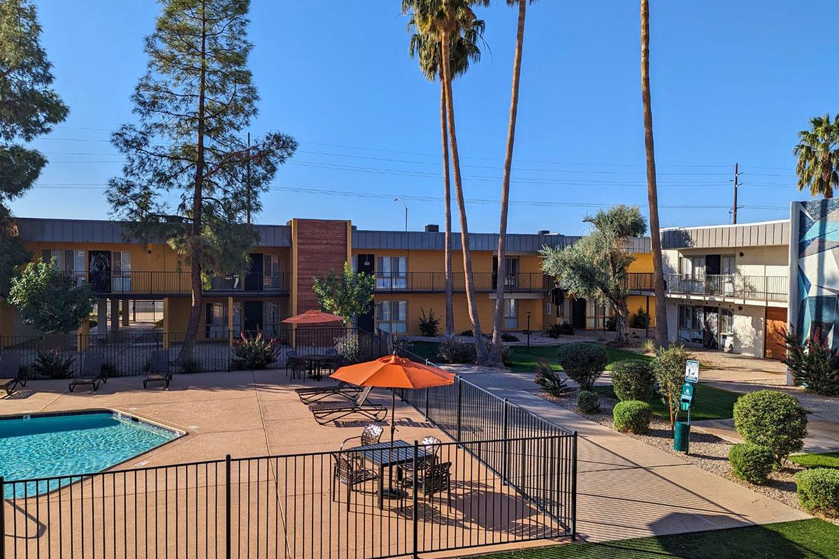 A sunny view of a residential area featuring a swimming pool surrounded by lounge chairs and an orange umbrella. Palm trees and shrubs are present, with two-story buildings in the background. The scene is bright and inviting, typical of a warm climate setting.