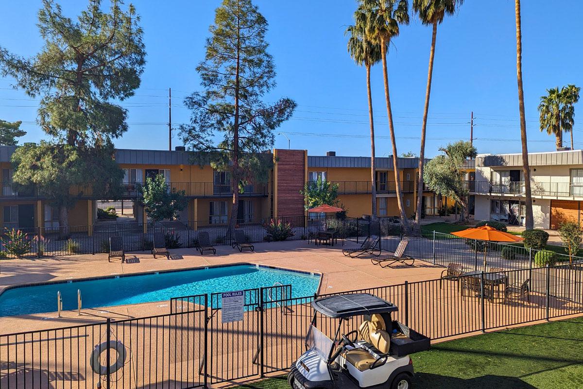 View of a hotel courtyard featuring a swimming pool surrounded by lounge chairs and umbrellas. Palm trees stand tall against a clear blue sky, with a golf cart parked nearby. The hotel buildings are two stories high, displaying a simple, colorful facade.