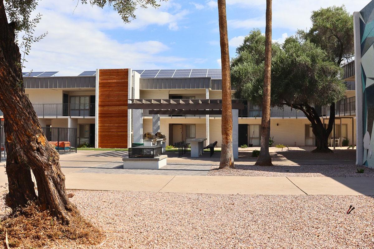 A modern multi-story building with solar panels on the roof, featuring a central courtyard with a BBQ area. Surrounding the building are palm trees and gravel landscaping, under a partly cloudy blue sky. A mural can be seen on the side of the building.