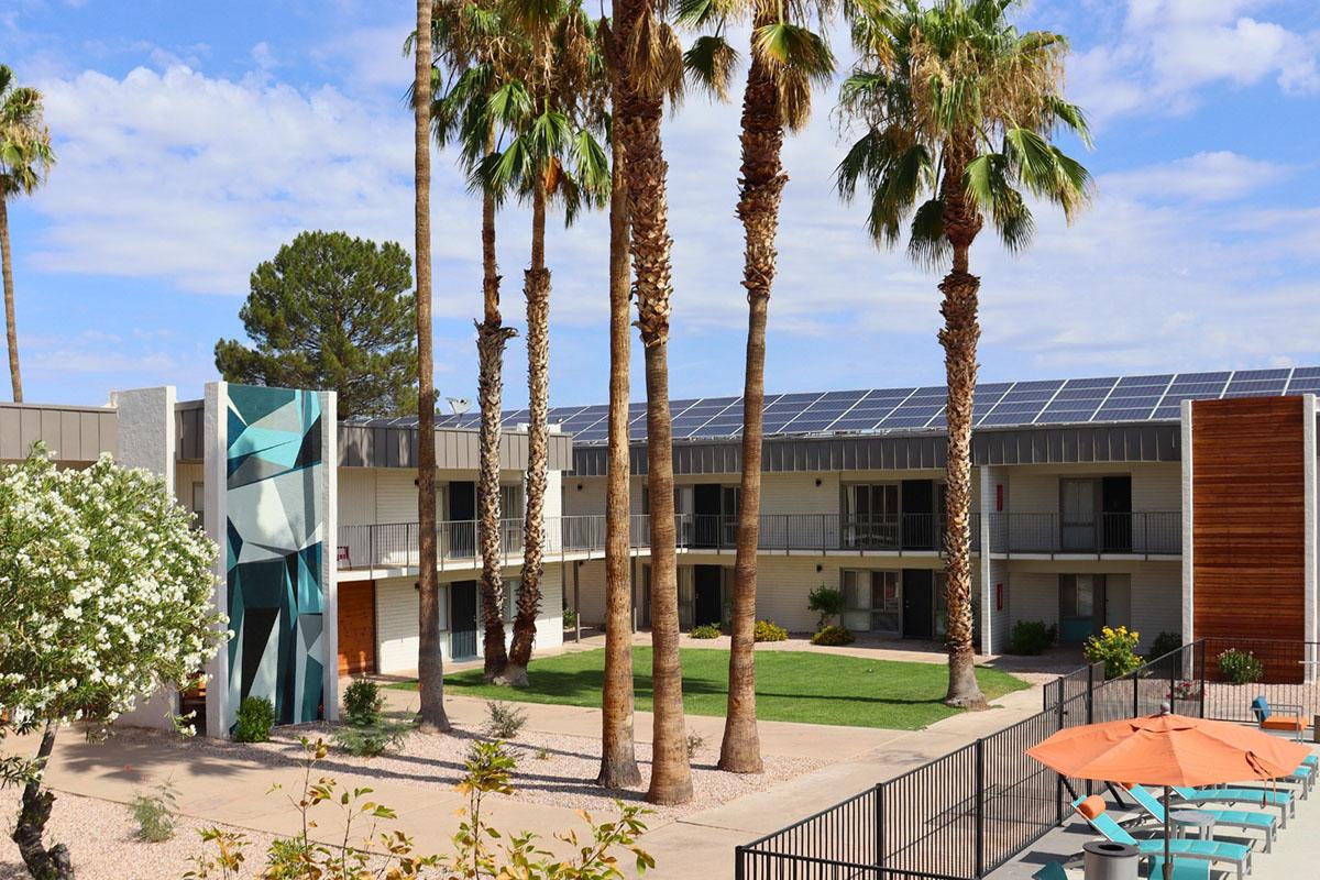 A view of a modern apartment complex featuring multiple stories, palm trees, and solar panels on the roof. The courtyard has green grass and well-maintained landscaping, with colorful outdoor furniture visible. The building's exterior showcases a contemporary design with a geometric mural on one side.