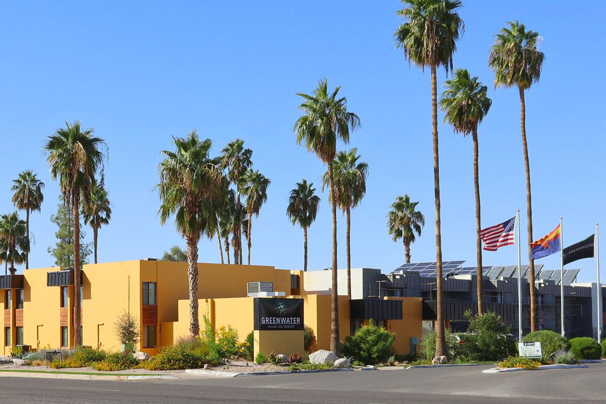A modern yellow building with palm trees in the foreground. The building displays a sign for "Greenwater." In the background, there are flags, including the American flag and a state flag. The area is well landscaped with shrubs and flowers, creating a vibrant, welcoming atmosphere.