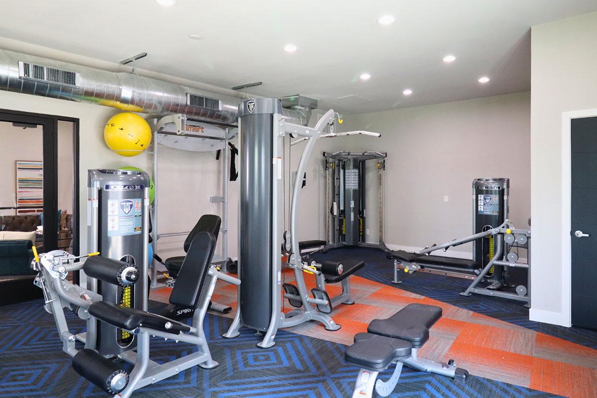 A modern gym interior featuring various exercise machines, including weightlifting stations, cable machines, and cardio equipment. The space has bright lighting and colorful flooring, creating an inviting atmosphere for workouts.