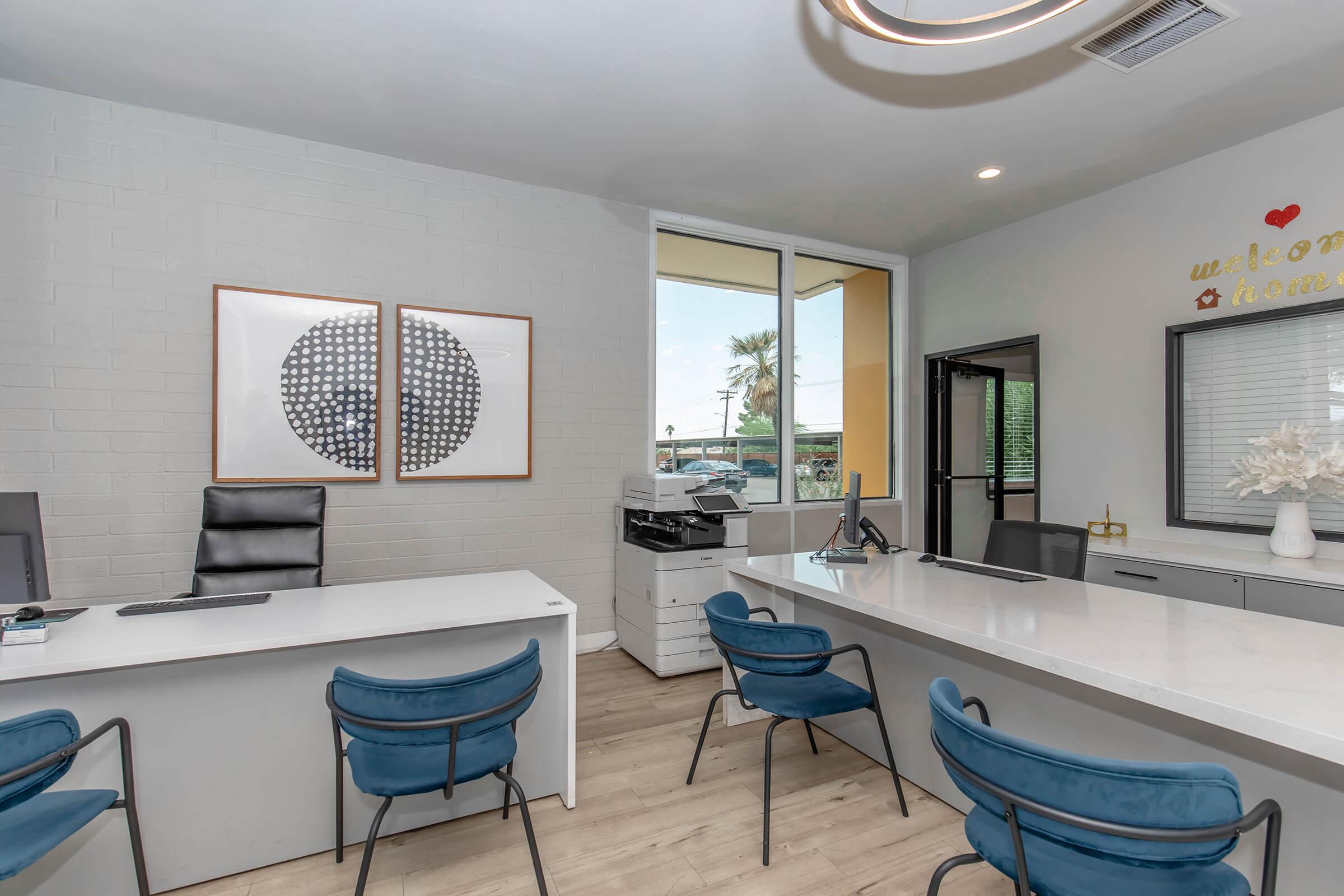 Modern office reception area featuring a sleek white desk, blue chairs, and artwork on the wall. Natural light streams through large windows, with views of palm trees outside. A printer and office supplies are visible, creating a welcoming atmosphere. Decorative elements include a "welcome home" sign.