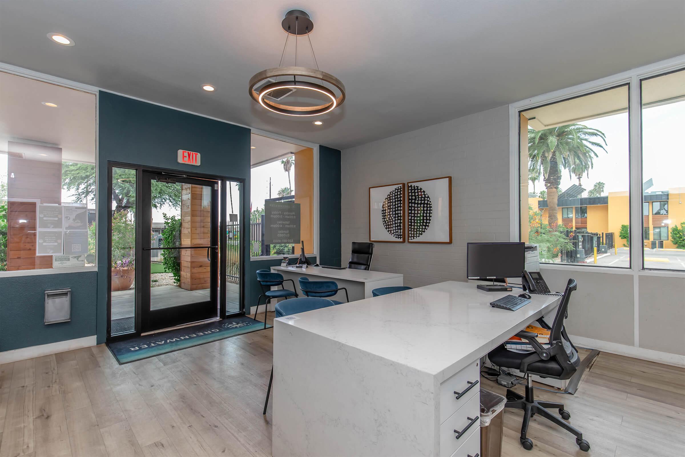 Brightly lit reception area featuring modern furnishings. The space includes a sleek white desk with chairs, a computer, and a welcoming entrance with glass doors. Decorative wall art with circular designs adds a contemporary touch, while large windows let in natural light and offer a view of palm trees outside.