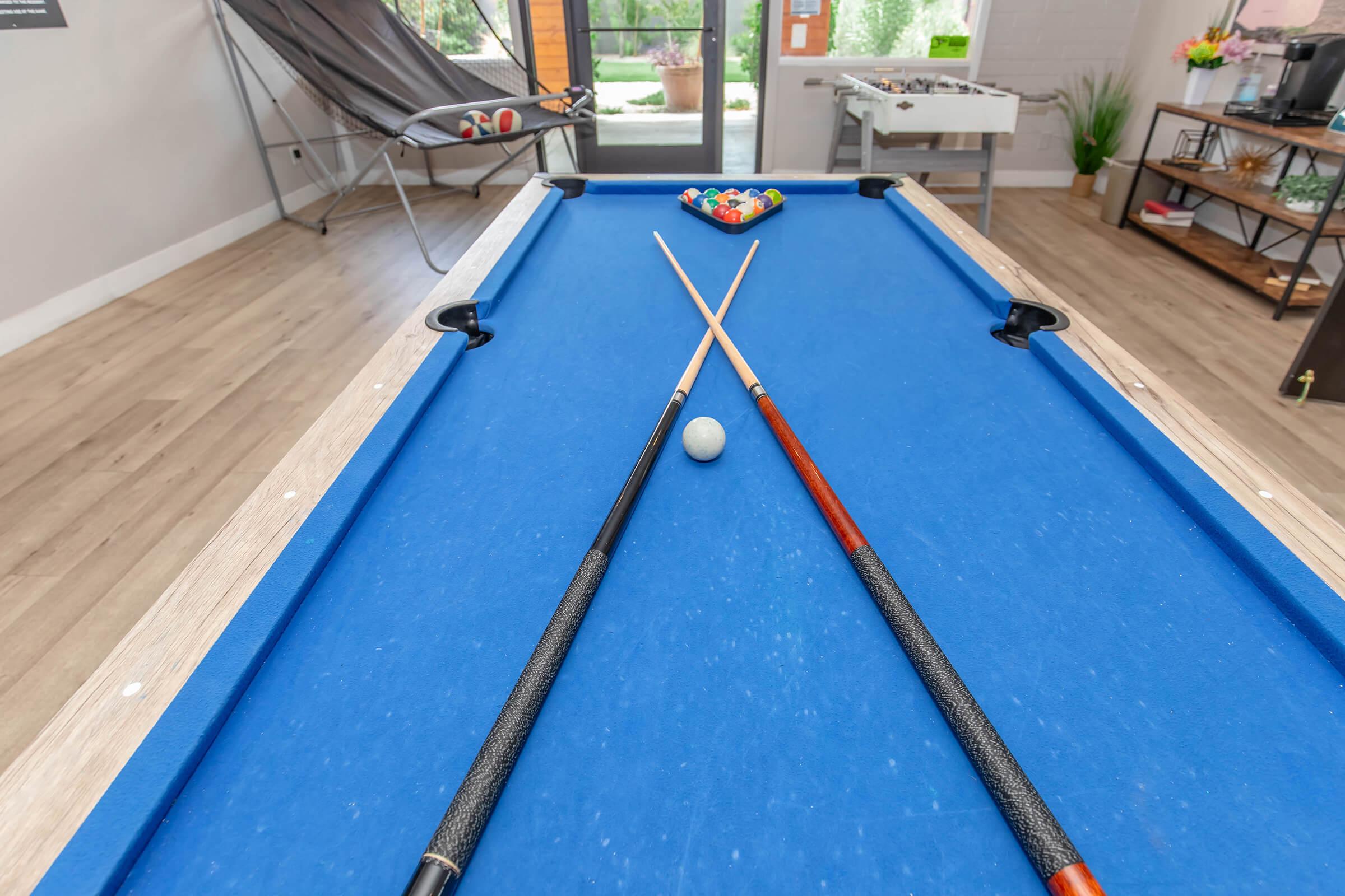 A blue pool table with two cue sticks crossed over a white billiard ball. In the background, a rack of colored balls waits for play. An open doorway leads to a bright outdoor space, and a lounge chair is visible to the side. The room features modern decor and natural lighting.