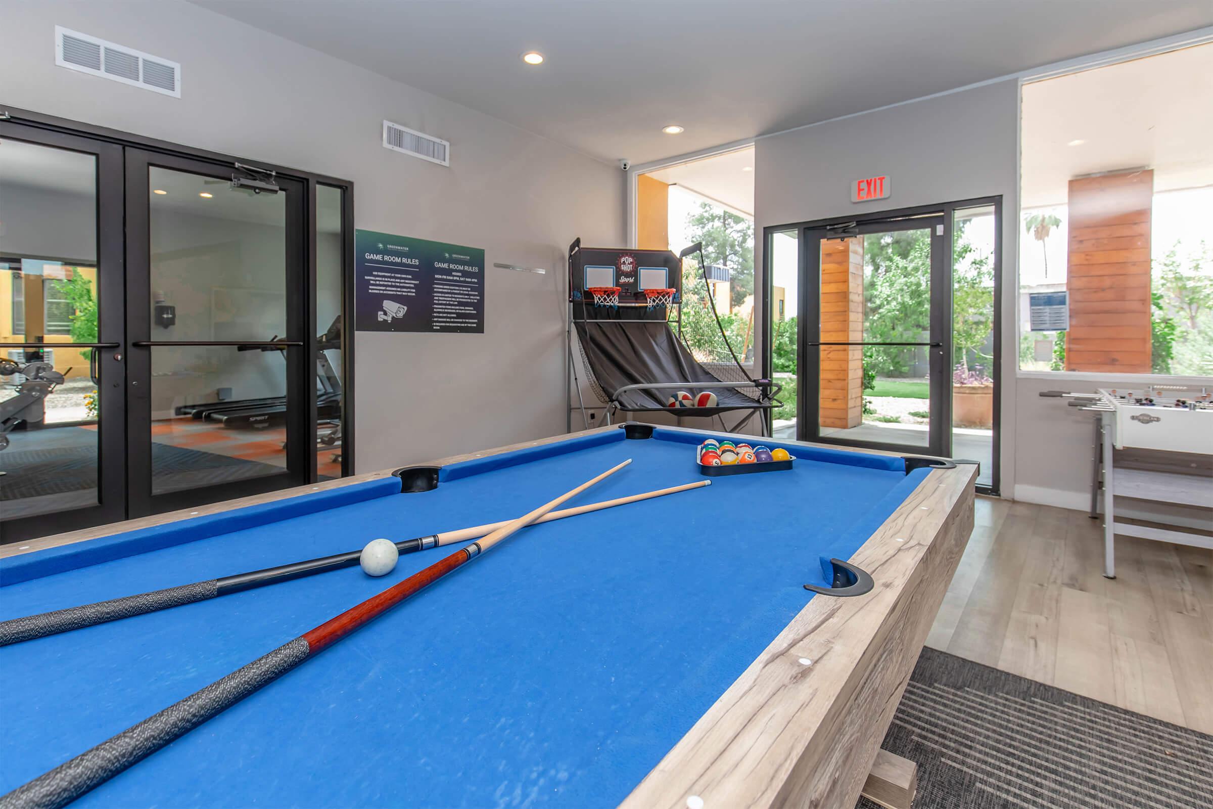 A well-lit game room featuring a blue pool table with two cues and a set of colorful billiard balls. In the background, a glass door opens to an outdoor area, and a ping pong table can be seen nearby. The walls are neutral-toned, enhancing the modern aesthetic of the space.