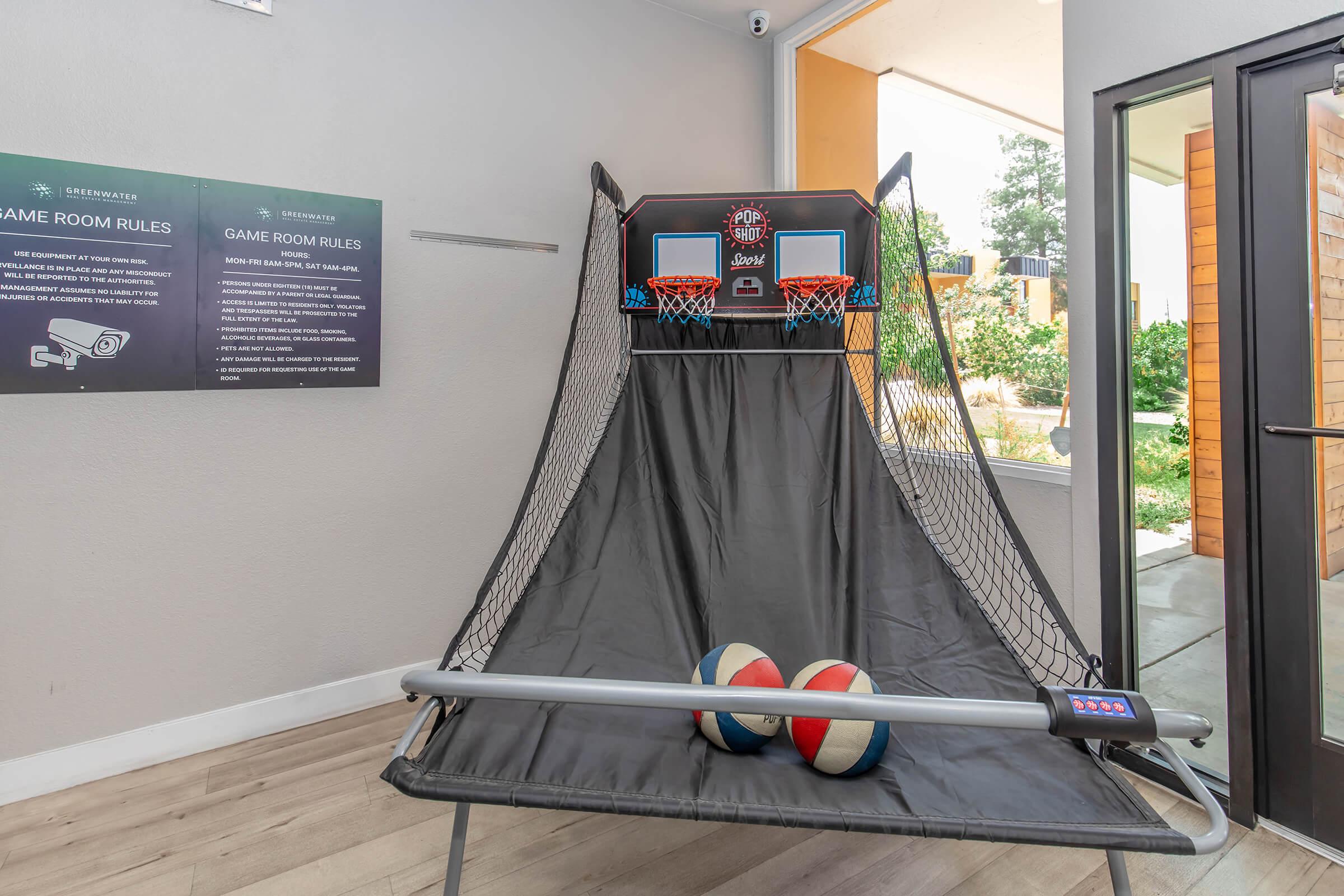 A modern indoor basketball shooting game with two hoops and a net, set against a wall. The game area includes two basketballs and a scoreboard. In the background, there are game room rules displayed on the wall, along with a bright window leading to an outdoor space.