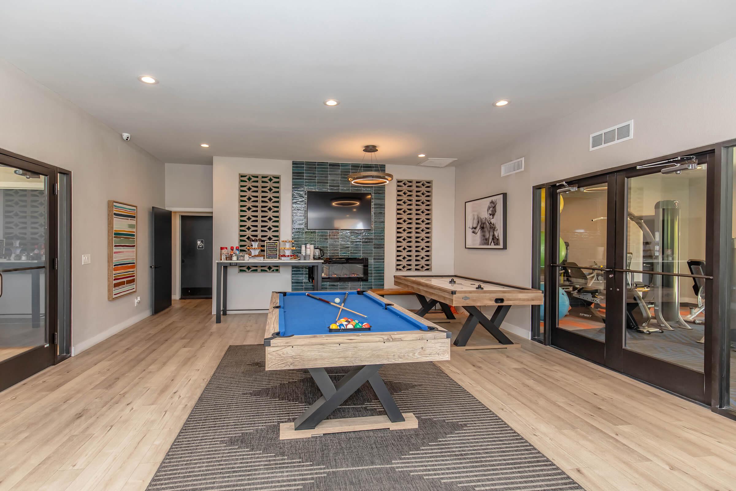 Indoor recreational area featuring a blue pool table, a wooden ping pong table, and a small bar station. Large windows for natural light, modern decor with vibrant art on the walls, and a fitness area visible in the background. The flooring is light wood, creating a warm and inviting atmosphere.