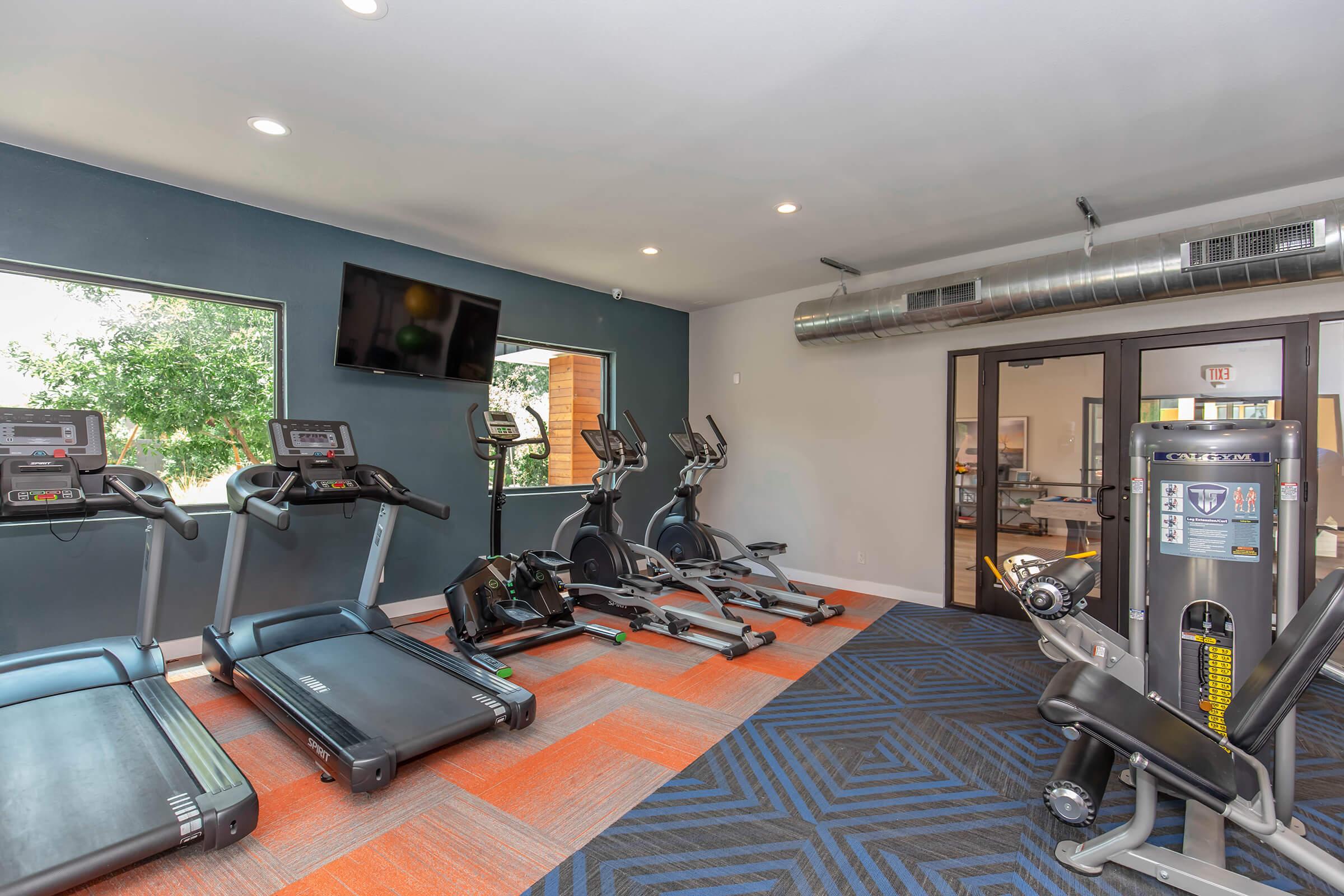 A modern gym featuring several exercise machines, including treadmills, elliptical trainers, and a weight machine. The space has a large window providing natural light, with blue and orange carpeting, and a wall-mounted television for entertainment during workouts.