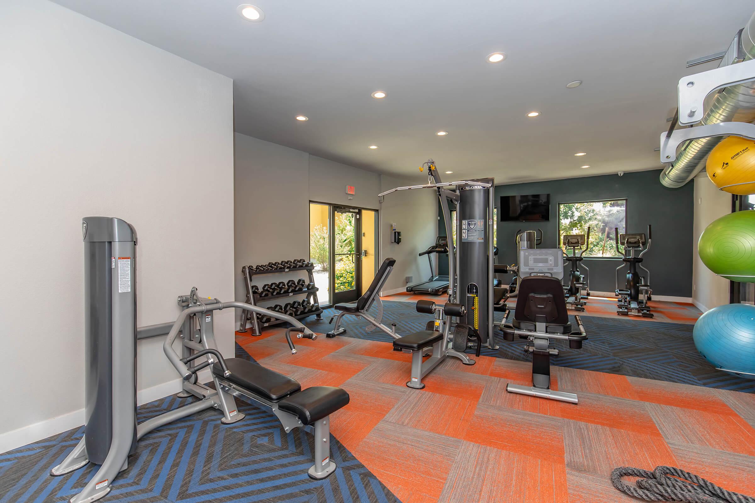 A modern gym interior featuring a variety of exercise equipment including weight machines, benches, dumbbells, and a cable machine. Large windows provide natural light, and exercise balls are arranged in one corner. The flooring has a geometric pattern with blue and orange colors.