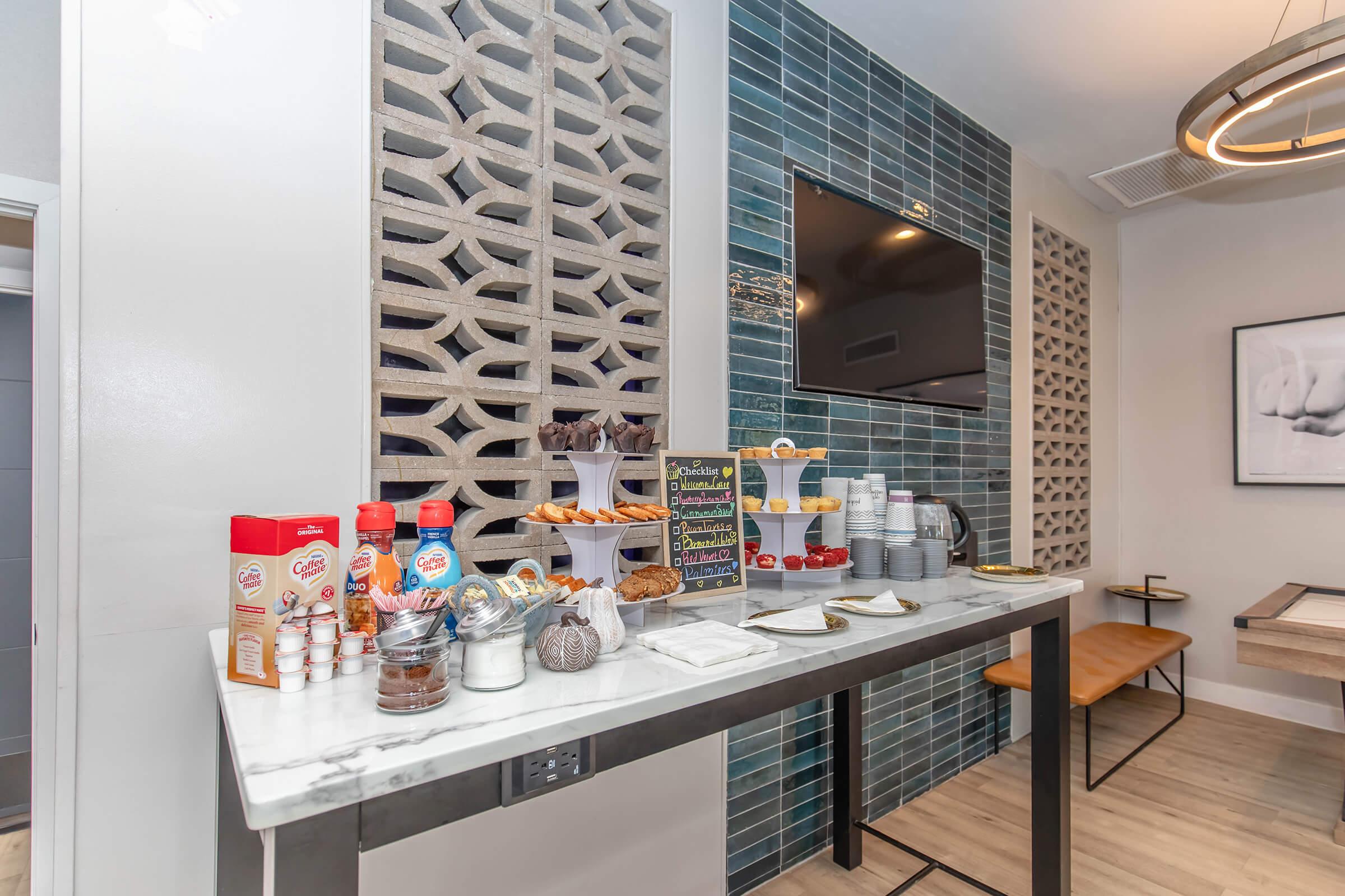 A decorated table with various breakfast items including pastries, snacks, and condiments. There are bottles of flavored coffee creamers, a coffee station with cups, and a menu board displaying offerings. The backdrop features decorative tile work and a mounted television.