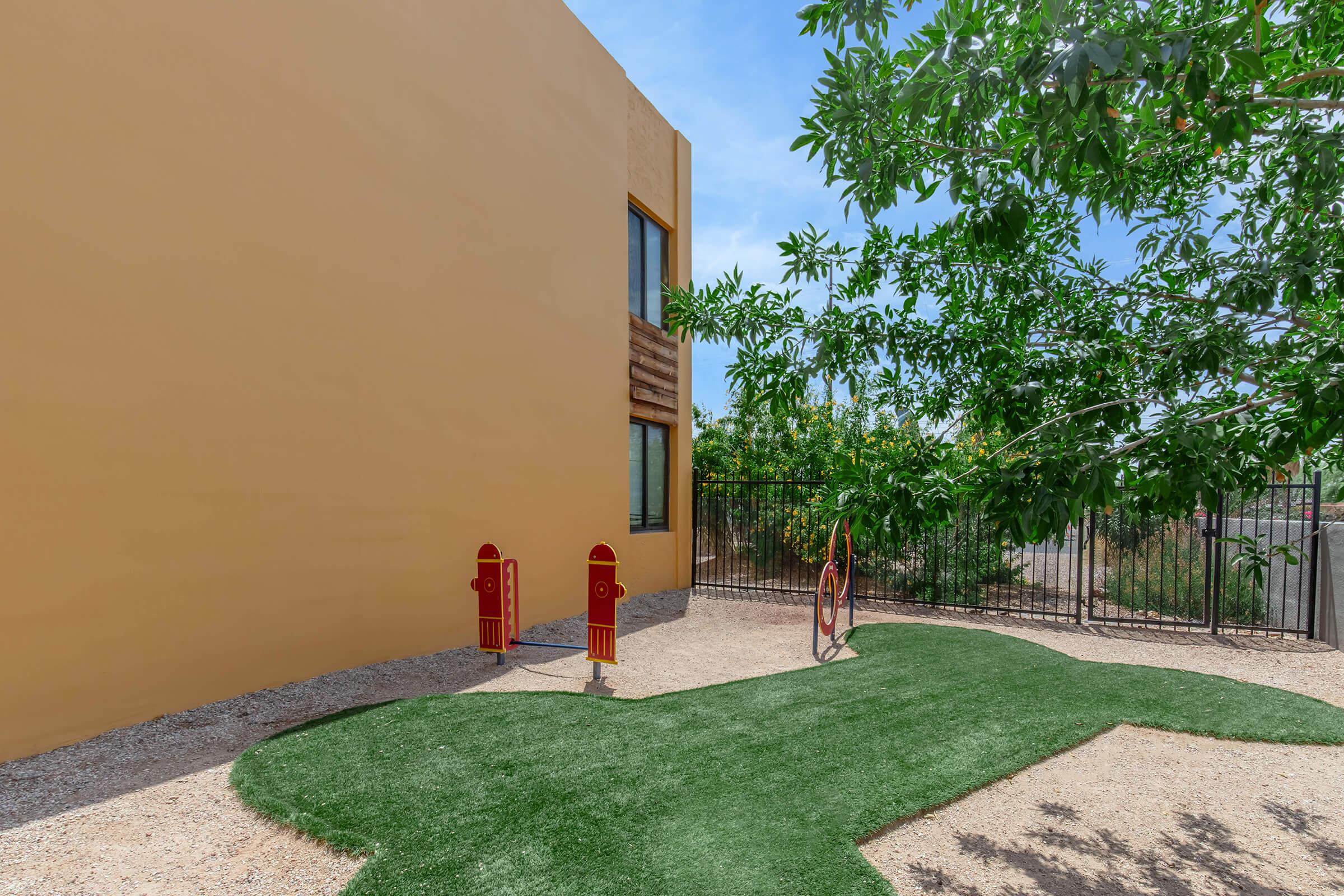 A small outdoor play area with artificial grass, featuring two colorful play structures resembling red fire hydrants. In the background, a yellow wall is partially visible, and there is a leafy green tree providing some shade. The scene is bright and inviting, perfect for young children to explore.