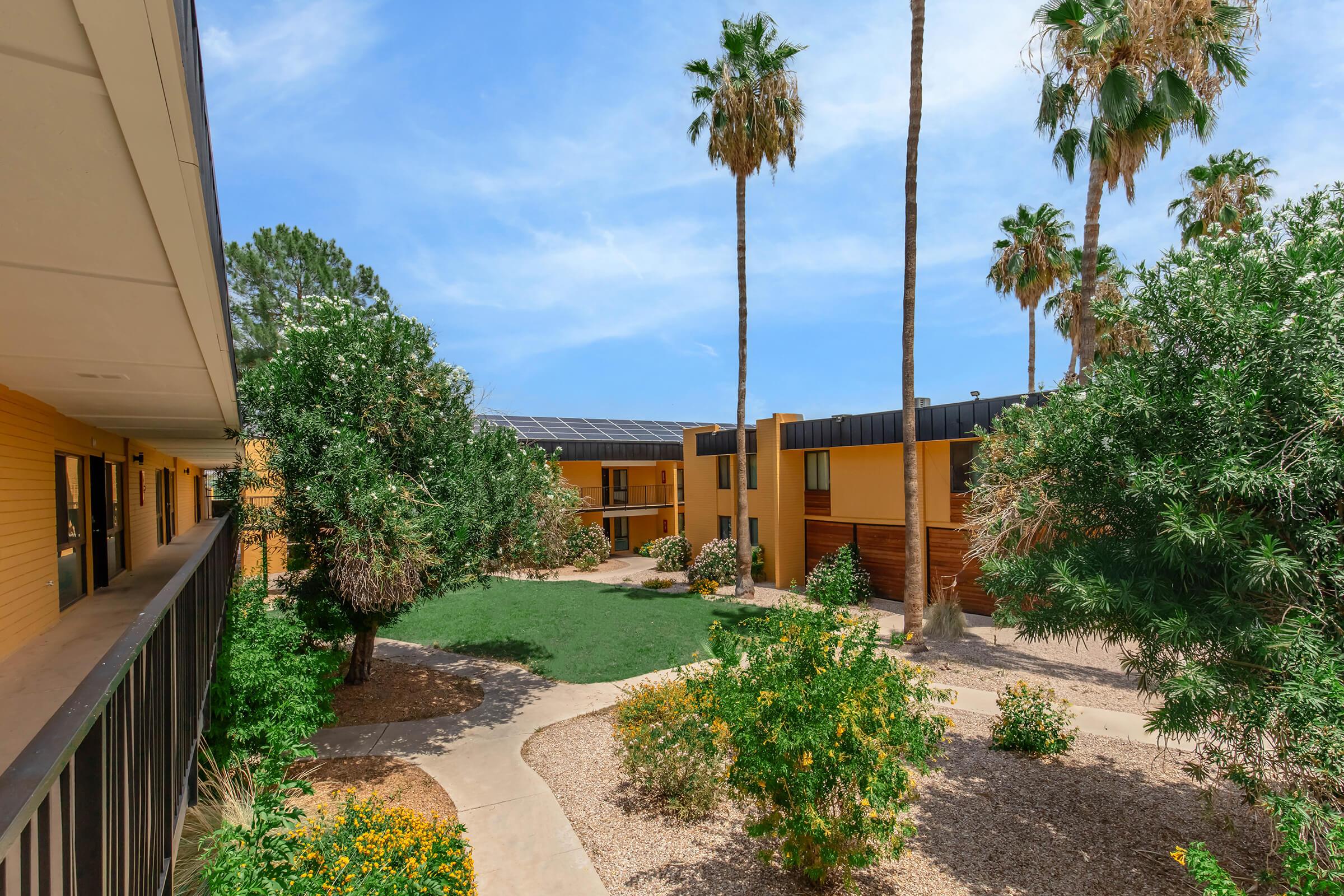 View of a landscaped courtyard surrounded by two-story buildings with yellow siding. Palm trees and various shrubs are visible, with a path winding through the greenery. The sky is bright blue, indicating sunny weather. Solar panels are on the roof of one of the buildings.