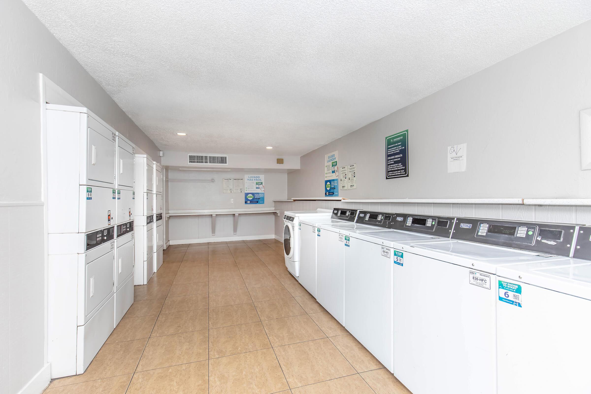 A clean, spacious laundry room with several white washing machines and dryers lined up against the walls. The floor is tiled, and the walls are painted light gray. Bright overhead lights illuminate the area, which includes a folding counter and informational signs on the walls.