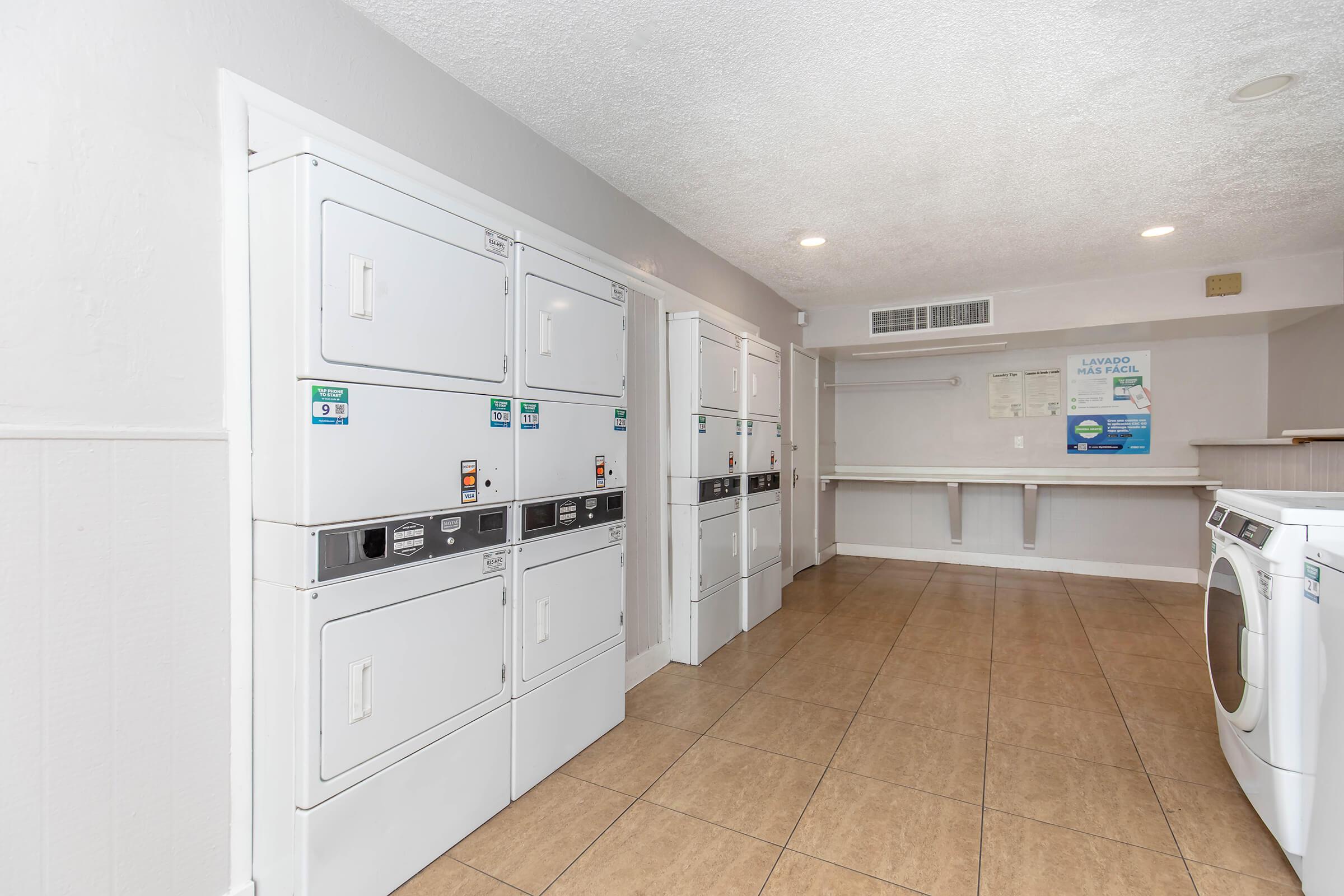 A clean and modern laundry room featuring several white coin-operated washers and dryers lined against the wall. The floor is tiled, and there's a folding table at the end of the room. The walls are painted in light colors, creating a bright and inviting atmosphere.