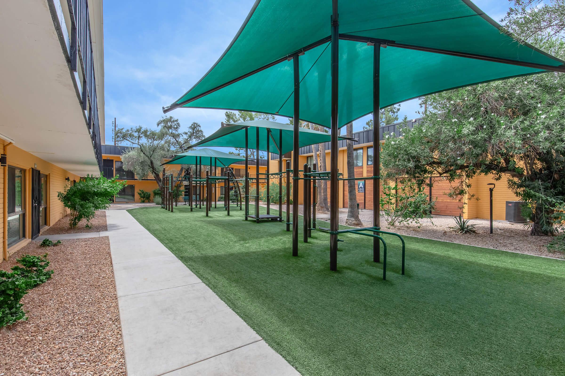 A landscaped outdoor area with artificial grass, featuring multiple green shade sails over swings and benches. Surrounding the space are shrubs and trees, with a pathway leading through the area. The setting appears to be part of an apartment complex or recreational facility.