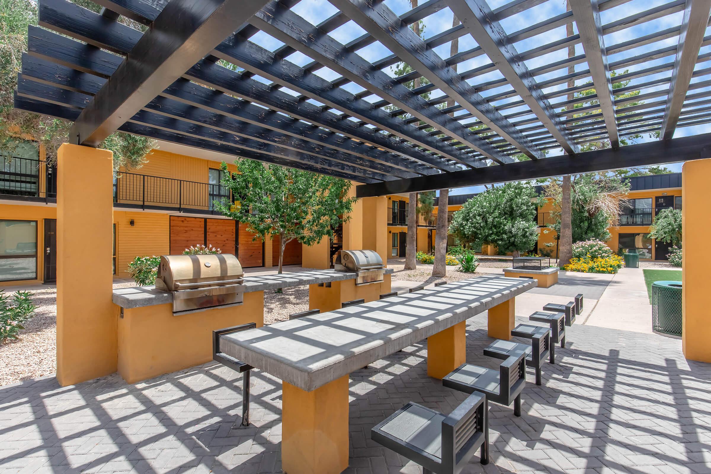 An outdoor grilling area featuring a large stone table with black metal chairs, under a wooden pergola. Two barbecue grills are positioned on the sides, surrounded by lush greenery and flowerbeds in a courtyard setting with yellow buildings in the background.