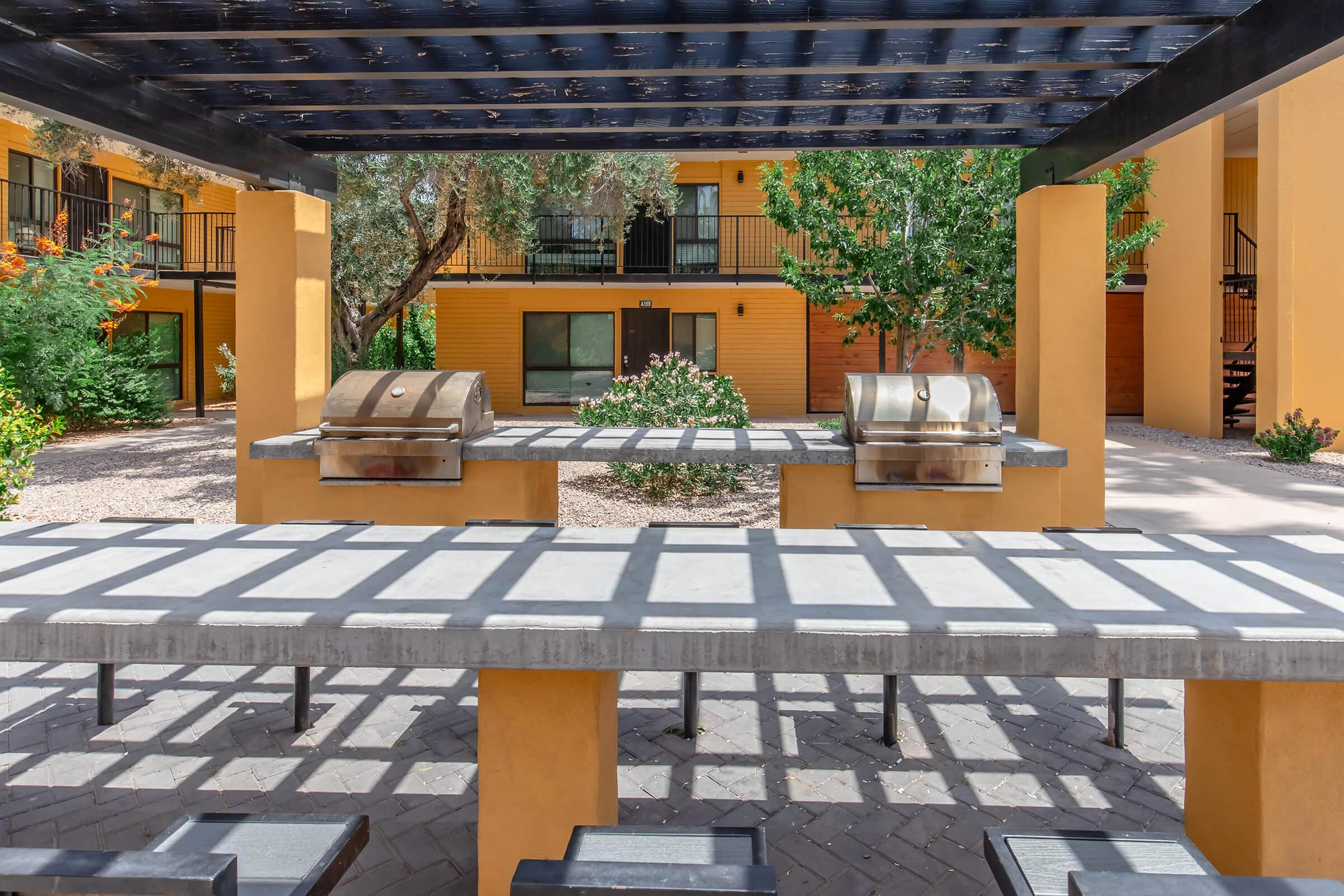 Outdoor grilling area featuring two stainless steel barbecue grills under a shaded pergola. A large concrete table with benches is situated in front, surrounded by landscaped greenery and an apartment building in the background, providing a communal space for gatherings.