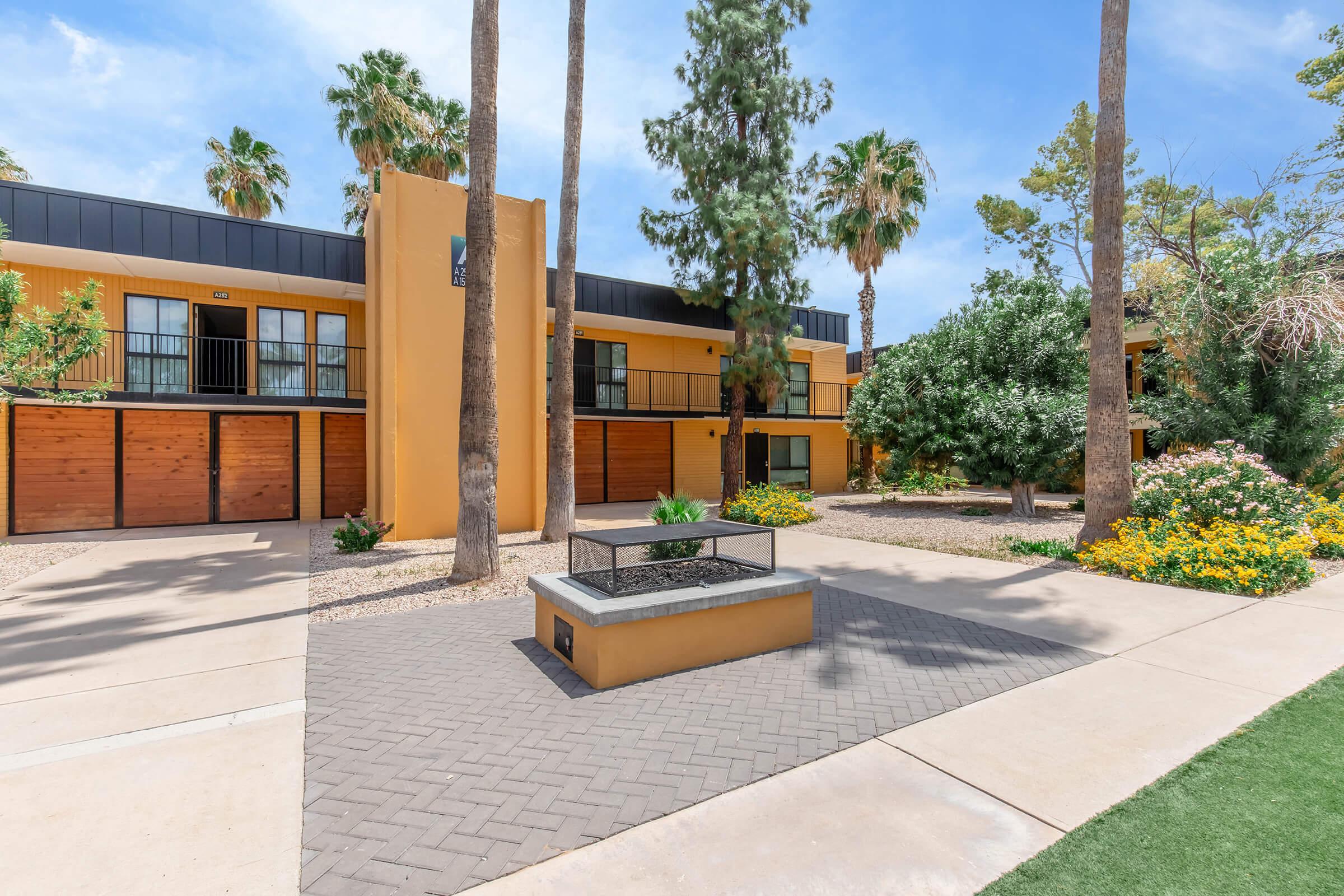 A sunny courtyard featuring modern apartment buildings with wooden garage doors. The buildings are surrounded by palm trees and lush greenery, complemented by colorful flowering shrubs, creating a vibrant outdoor space. A central fire pit adds a cozy element to the area.