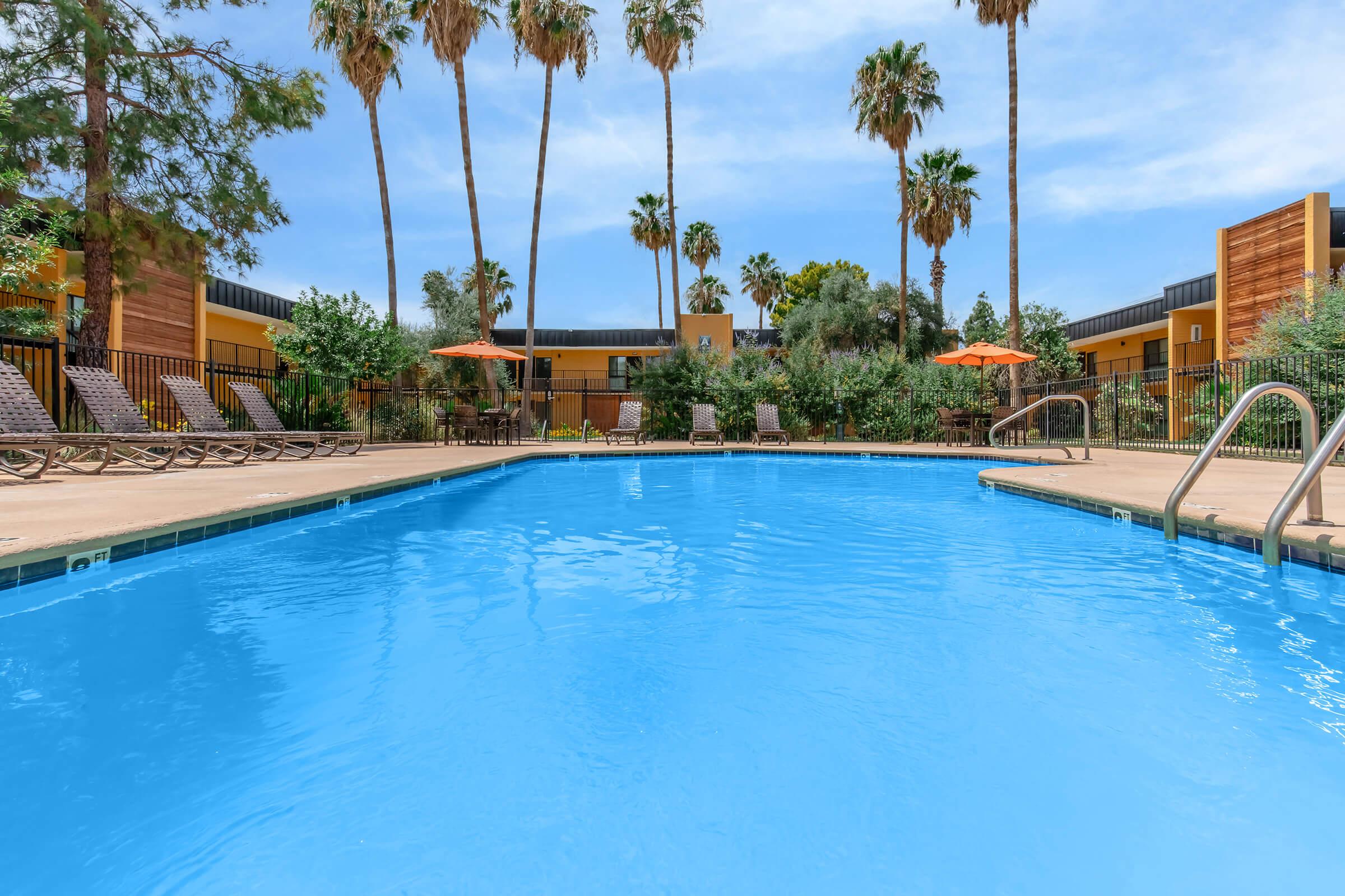 Clear blue swimming pool surrounded by lounge chairs and palm trees, with orange umbrellas providing shade. In the background, modern buildings with a warm color palette are visible against a partly cloudy blue sky. The scene evokes a relaxing outdoor atmosphere.