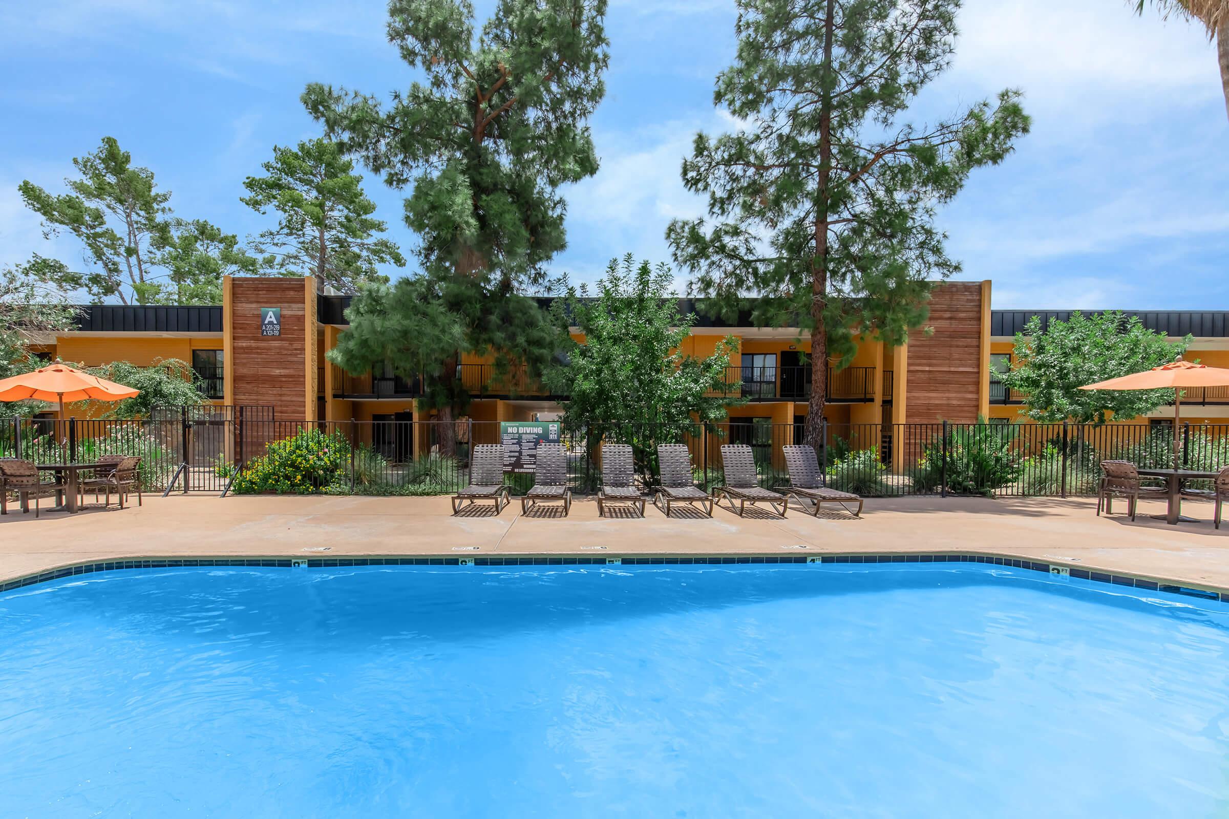 A bright outdoor pool surrounded by lounge chairs and orange umbrellas. In the background, there are lush green trees and a building with a wooden exterior. The sky is partly cloudy, creating a vibrant and inviting atmosphere.