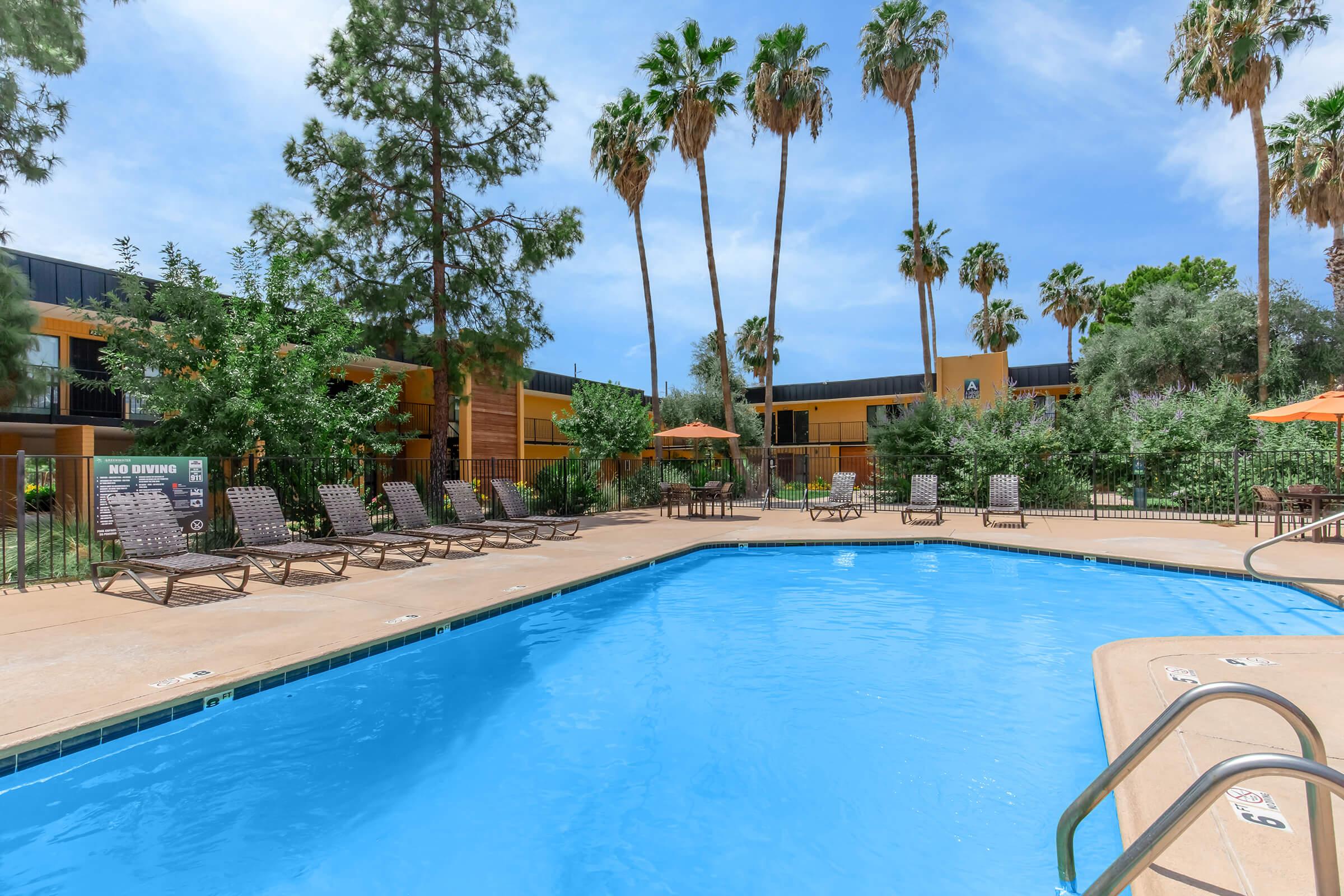 A resort-style swimming pool surrounded by lounge chairs and palm trees. Orange umbrellas are placed near the pool area, with a backdrop of two-story buildings and lush greenery under a clear blue sky.