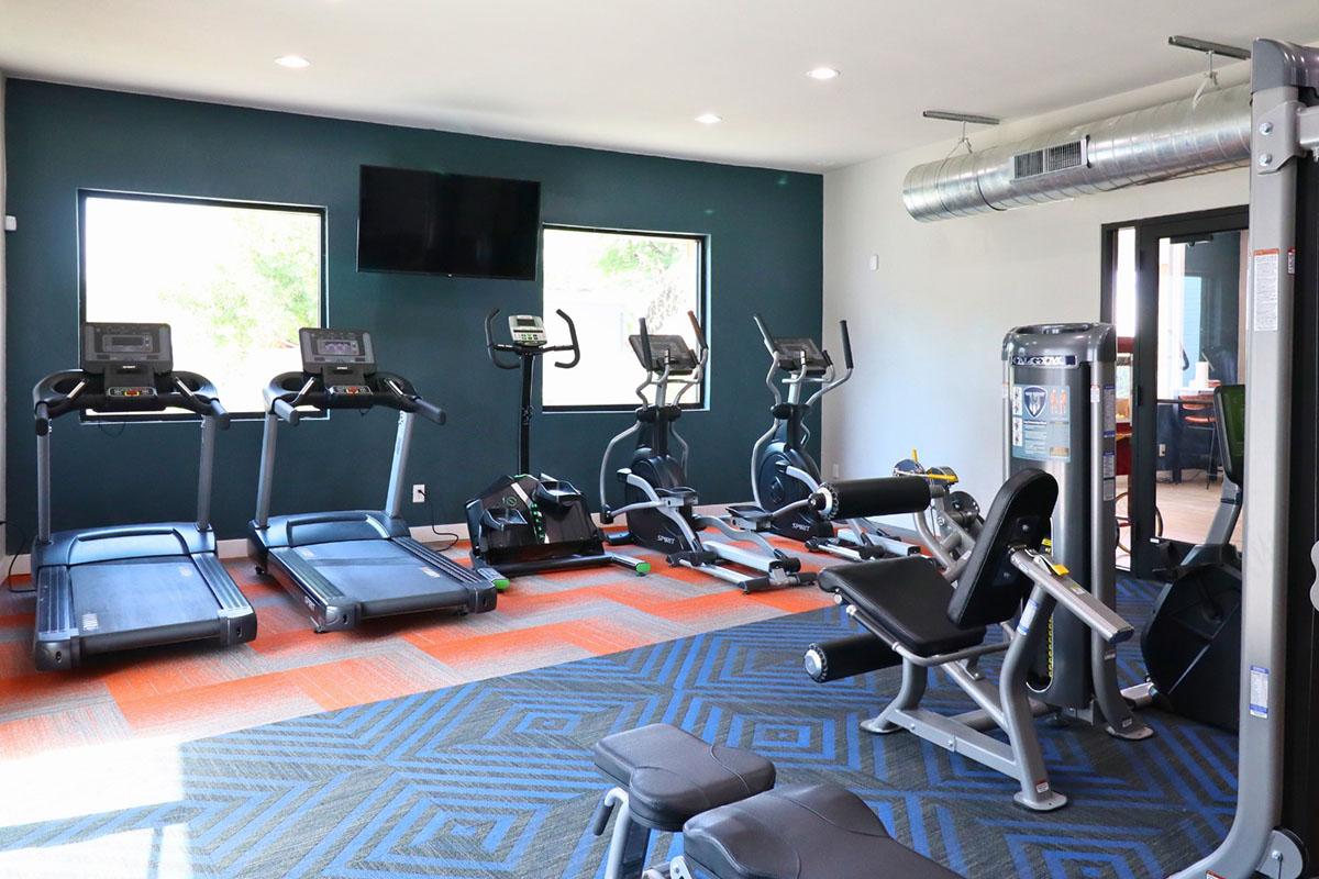 A modern gym interior featuring several exercise machines including treadmills, elliptical machines, and a multi-gym station. The space is well-lit with large windows and has a vibrant floor design. A TV is mounted on the wall, and there are weights on the floor.