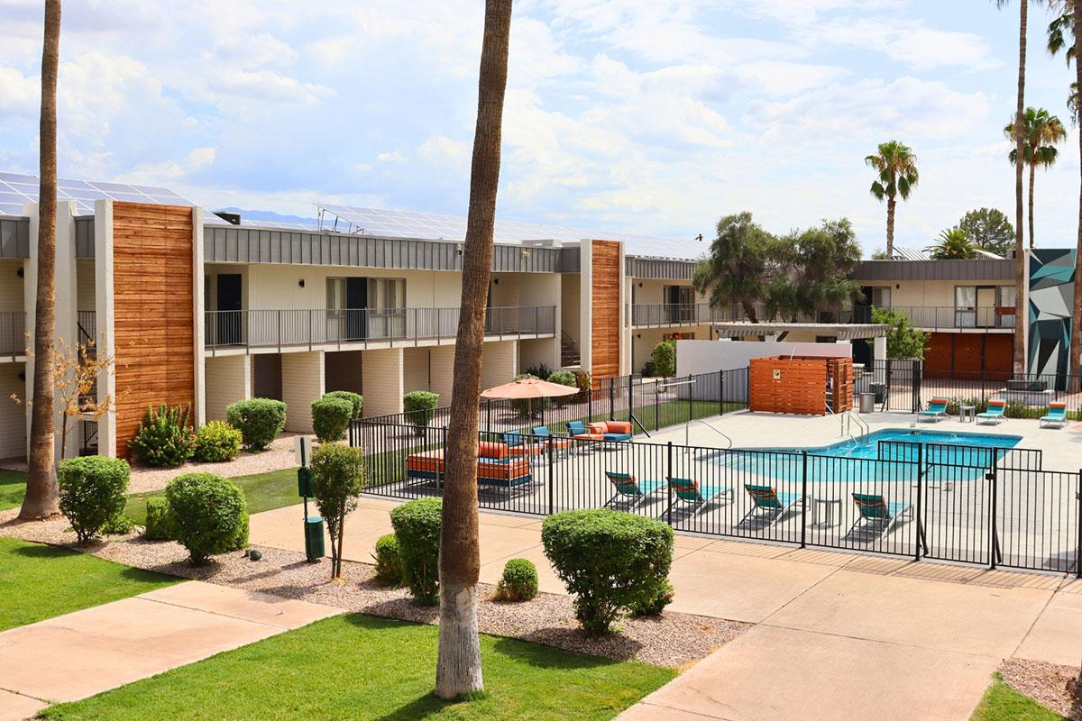 A view of a hotel courtyard featuring a swimming pool surrounded by lounge chairs and umbrellas. The building has a modern design with a mix of wood and metal finishes, palm trees, and well-maintained landscaping. Clear skies above enhance the inviting atmosphere of the outdoor area.