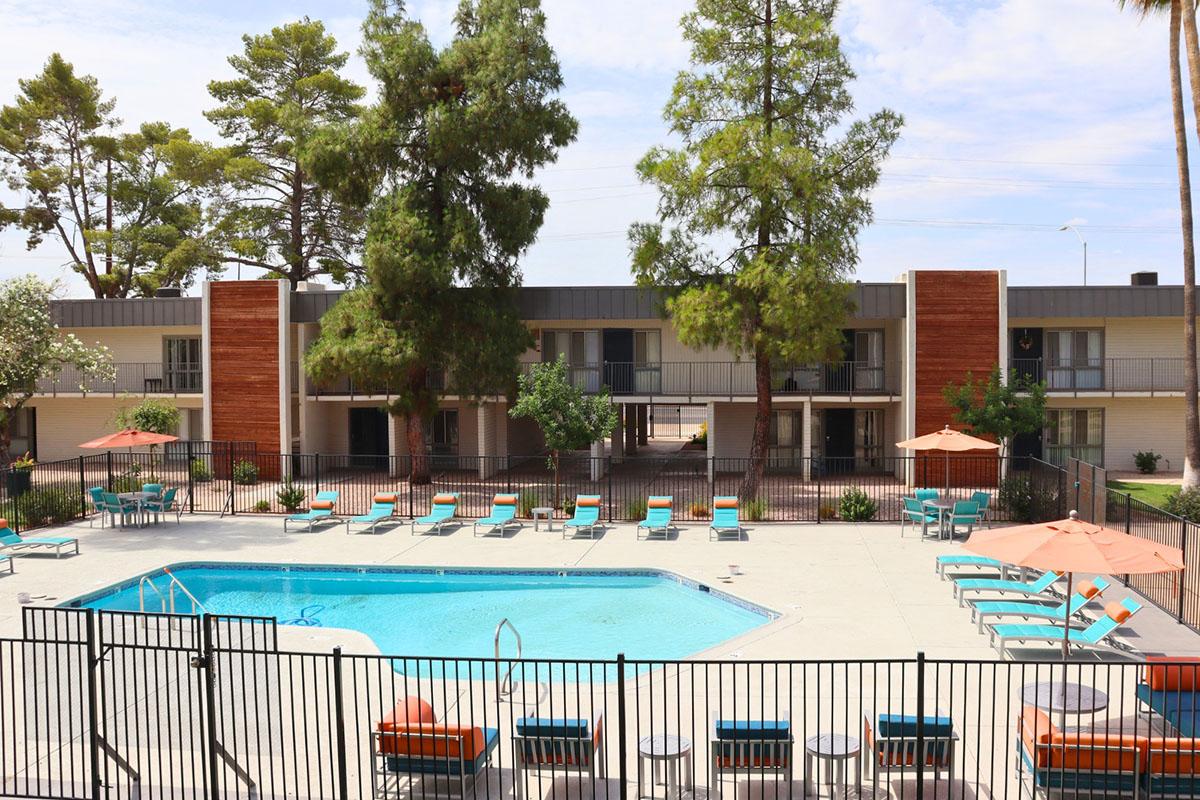 A clear view of a swimming pool surrounded by lounge chairs and umbrellas, with a backdrop of two-story buildings and trees. The pool area is fenced, featuring a sunny environment with a few clouds in the sky.