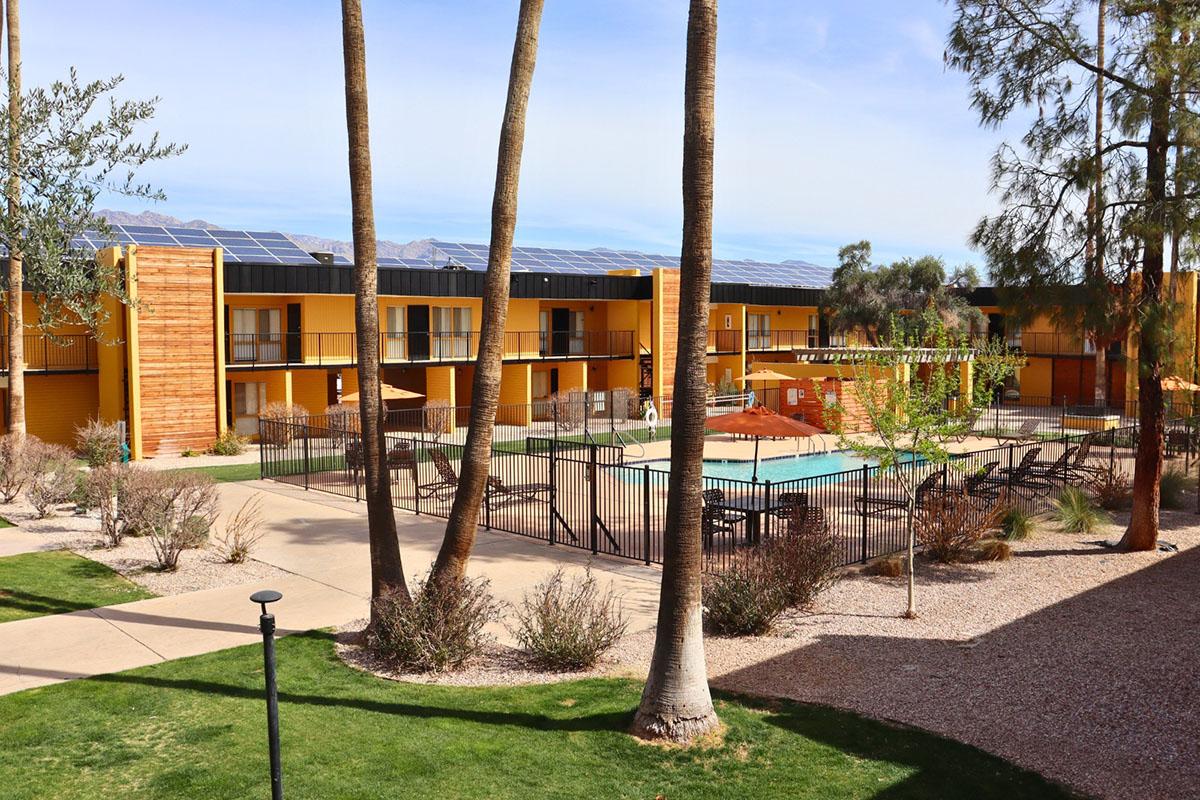 A scenic view of a modern hotel courtyard featuring a swimming pool, surrounded by palm trees and desert landscaping. The hotel buildings are painted in bright yellow and have solar panels on the roofs. Blue skies and light clouds are visible in the background.