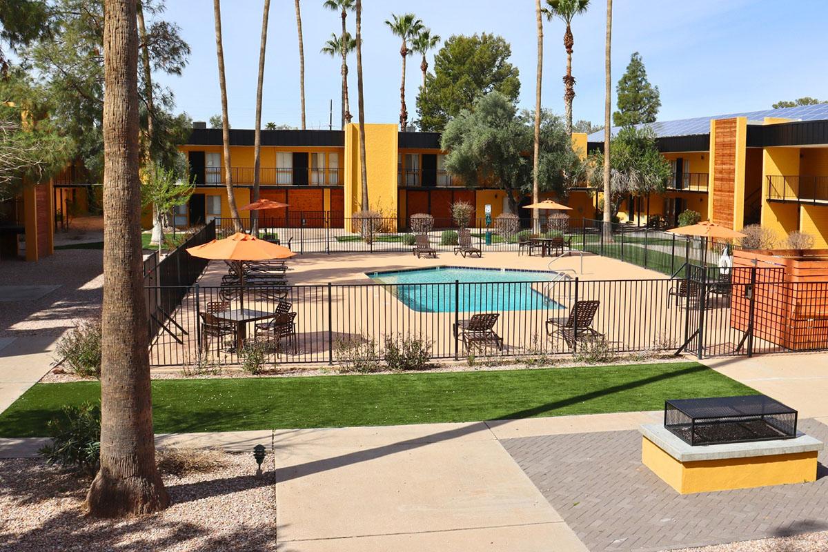 A view of a pool area surrounded by lounge chairs and orange umbrellas, enclosed by a black fence. Palm trees line the space, with yellow buildings in the background. The ground features a mix of concrete and grassy areas, creating a welcoming atmosphere for relaxation.