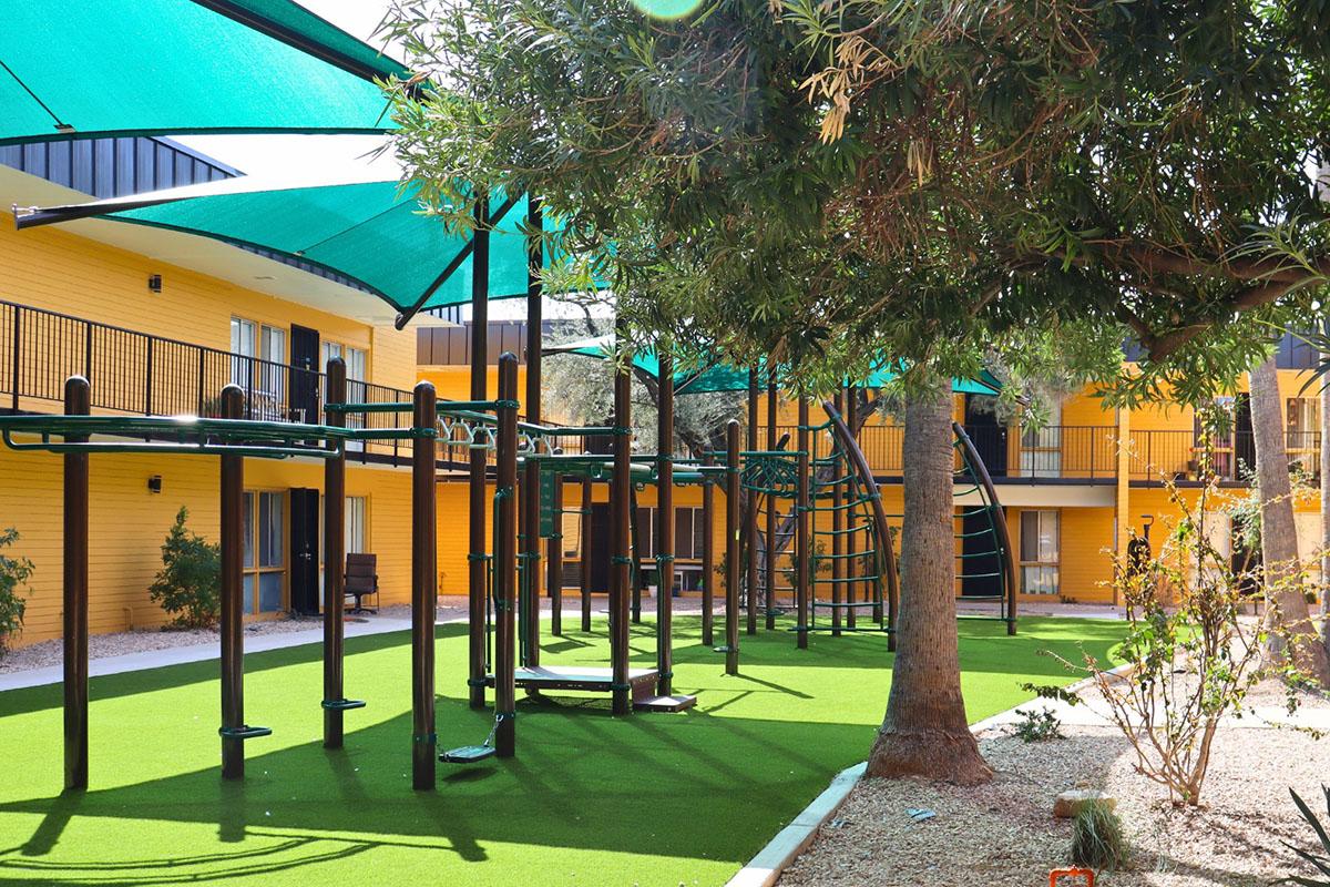 A vibrant outdoor play area featuring green artificial turf, playground equipment with climbing structures, and large shaded canopies. Surrounding the play area are small trees and shrubs, with bright yellow buildings visible in the background, creating a cheerful and inviting atmosphere.
