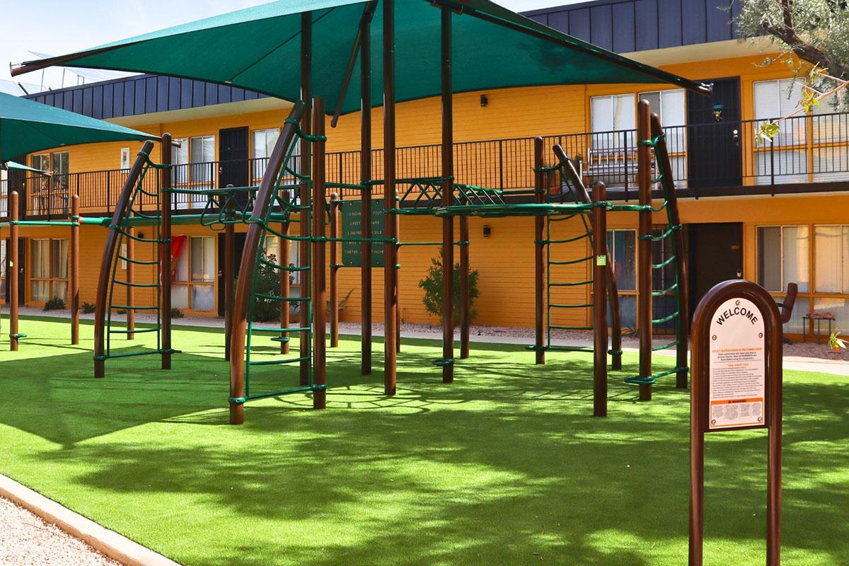 A playground with climbing structures and green shade canopies on a grassy area, set against the backdrop of a brightly colored building with balconies. A sign nearby reads "Welcome." The scene is inviting and designed for recreational activities.