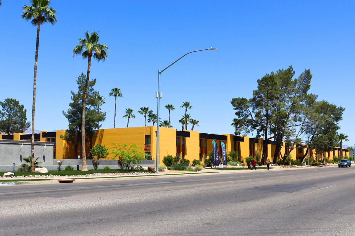 A street view of a sunny residential area featuring a row of yellow buildings with large windows, surrounded by palm trees and landscaped gardens. The scene includes a clear blue sky and a few pedestrians walking along the sidewalk.