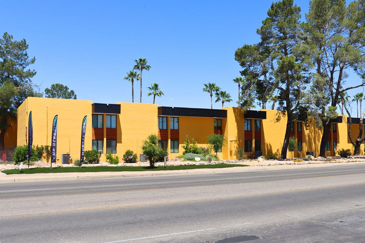 A bright yellow building with multiple windows and palm trees nearby. The building has decorative flags in front, and there is a clear blue sky above. The scene is set along a road with desert landscaping, creating a vibrant and inviting atmosphere.