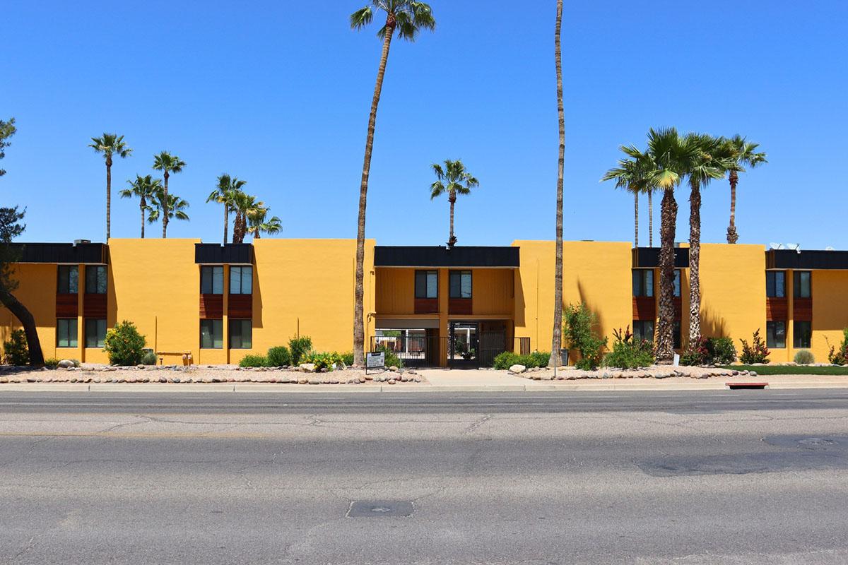 A bright yellow building with modern design features and large windows, flanked by tall palm trees. The view is from across the street, showing a clear blue sky above. The entrance is visible, along with landscaped areas in front of the building.