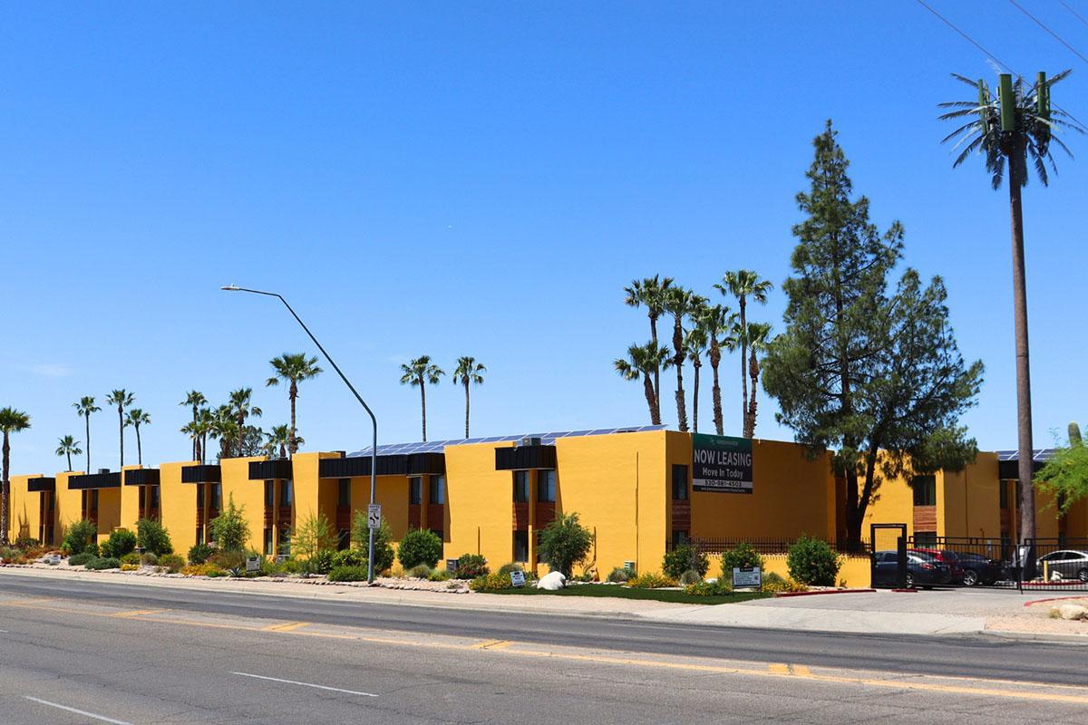 A bright yellow building with a "Now Leasing" sign, lined with palm trees and shrubs, sits alongside a street under a clear blue sky. The building features solar panels on the roof and is set in a sunny, open area.
