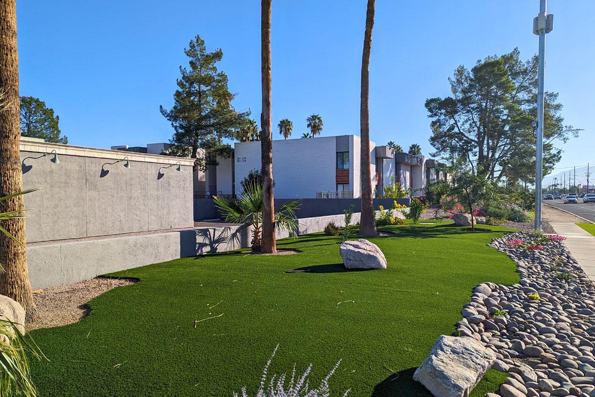 A landscaped area featuring green artificial turf, decorative rocks, and various plants. In the background, there are modern buildings surrounded by palm trees under a clear blue sky. The scene suggests a well-maintained outdoor space along a street.