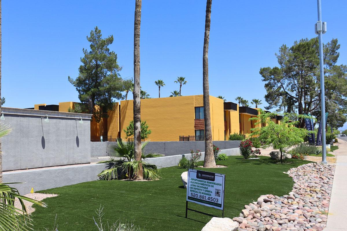 A view of a yellow building surrounded by palm trees and landscaped greenery, including artificial grass and rocky borders. A for-sale sign is visible in front, indicating a property available for purchase. The scene is set under a clear blue sky.
