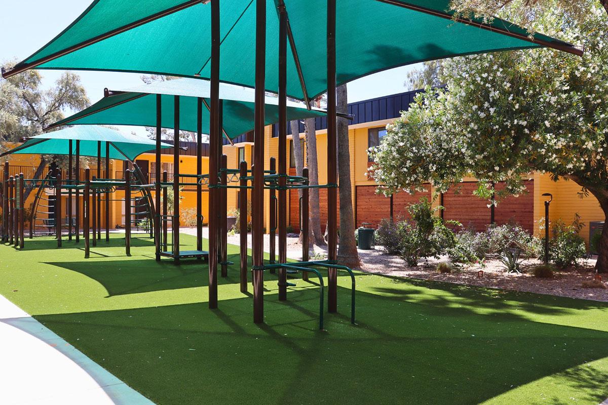 A bright outdoor playground featuring green artificial grass and shade structures. Various climbing and play equipment is visible, along with colorful yellow buildings in the background and lush greenery surrounding the area.